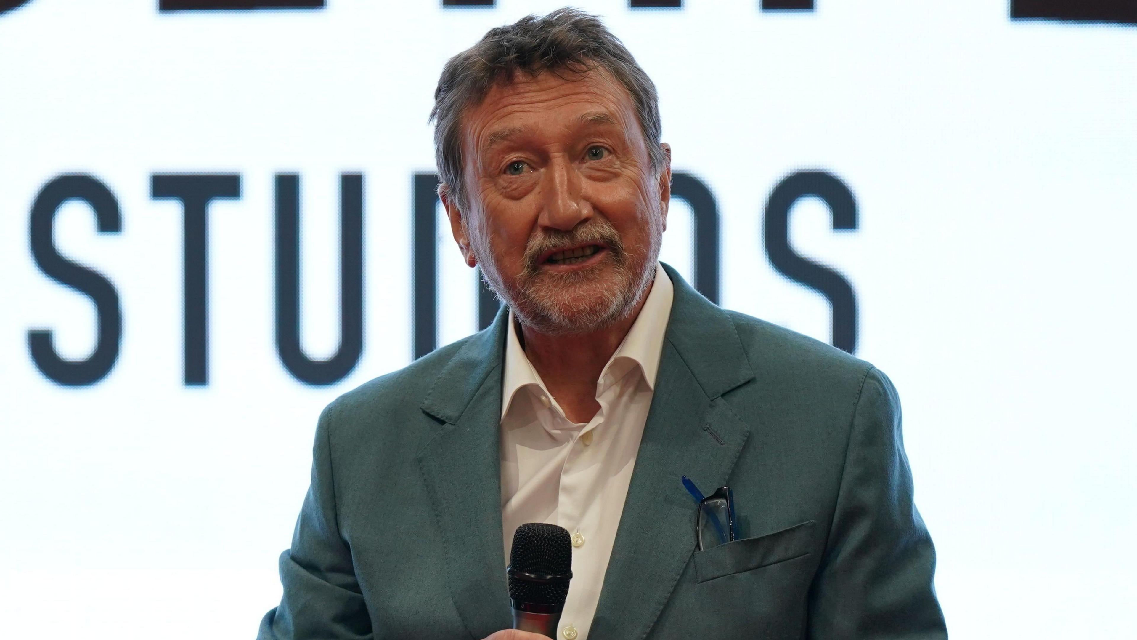 A man in a grey suit jacket and white shirt holds a microphone mid-speech. He is standing in front of a white backdrop with the word studios in capital letters. He has glasses tucked in his breast pocket, grey/brown hair and a neat greying beard.