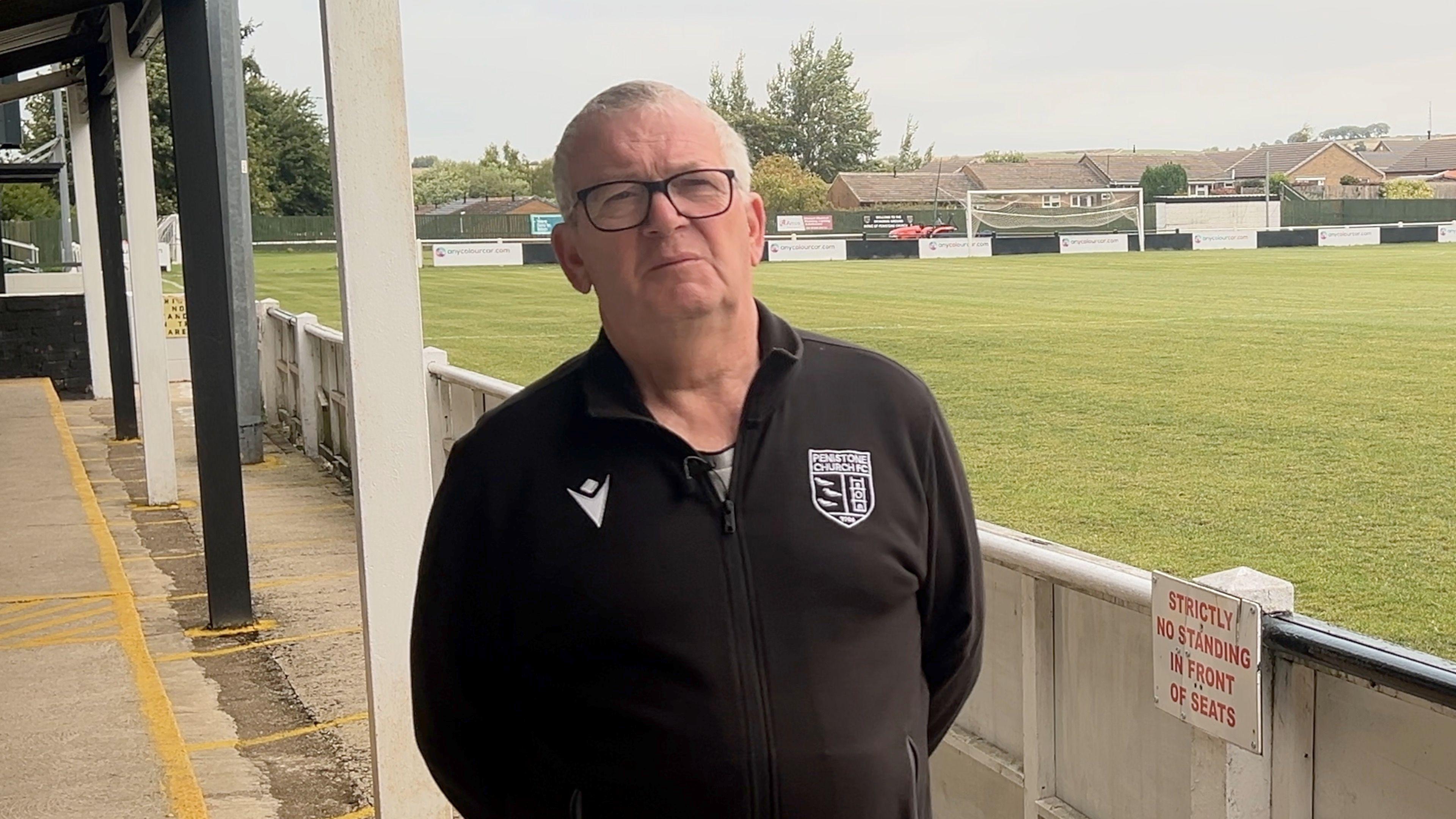 Dave Hampshire from Penistone Church FC stands in the stand of the club's stadium. He's wearing a club fleece and has glasses.