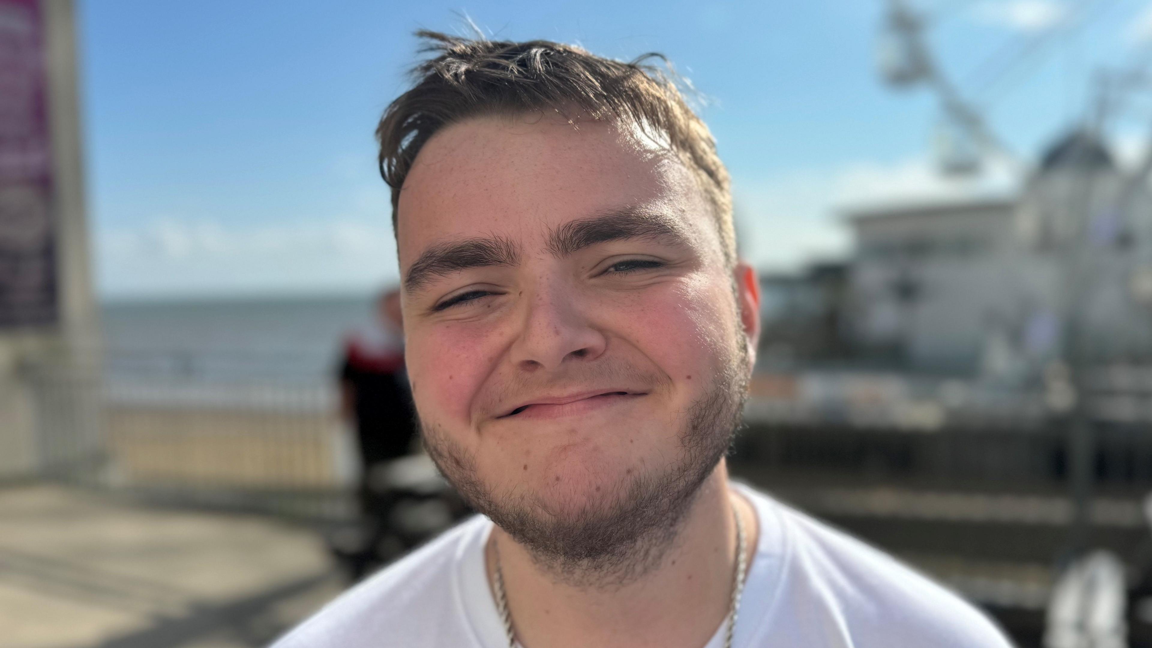 A smiling man wearing a white T-shirt with a chain around his neck. A beach is in the background.