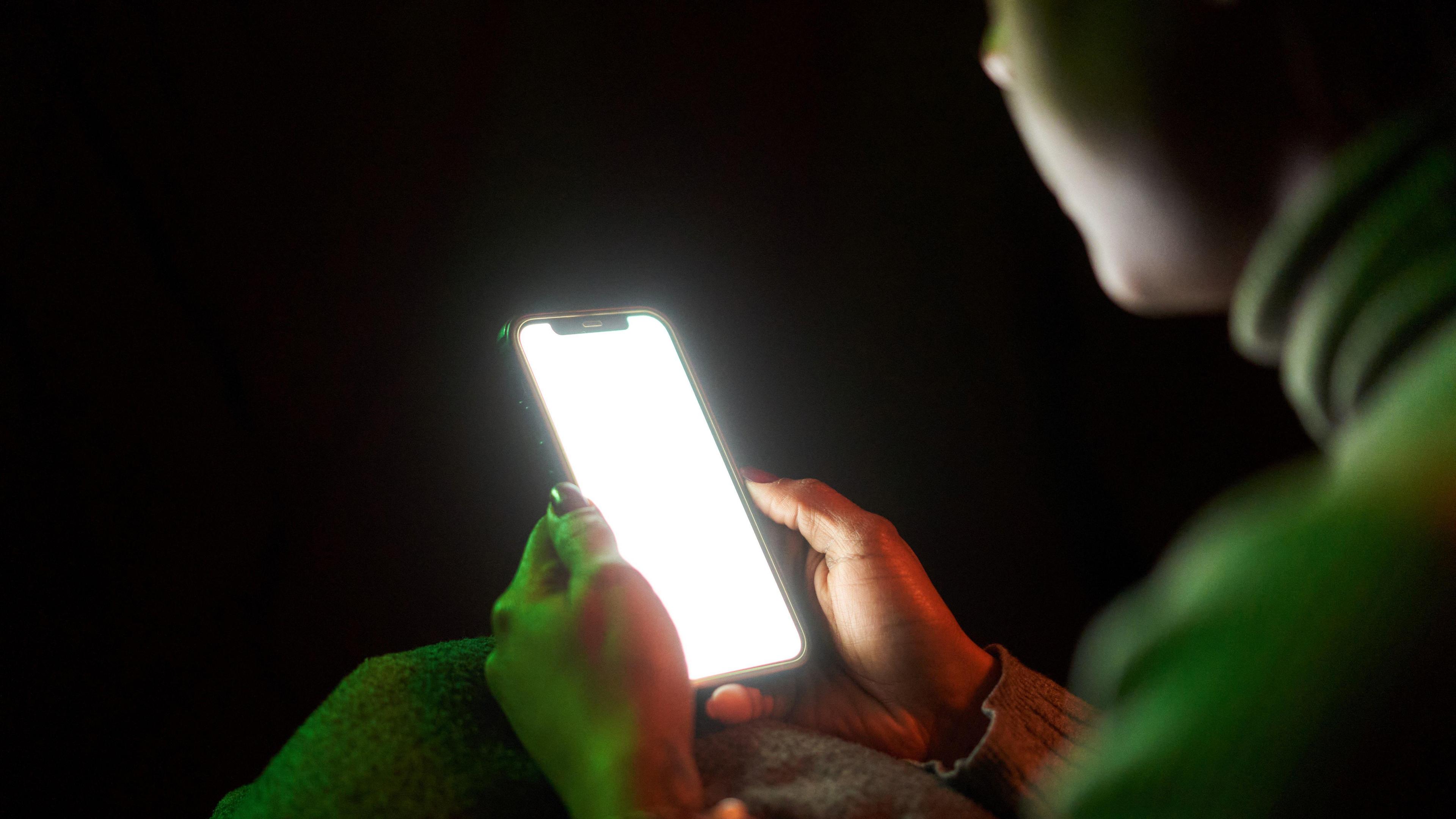 A woman sits in the dark holding a mobile phone. The light of the screen is partially illuminating her face. 