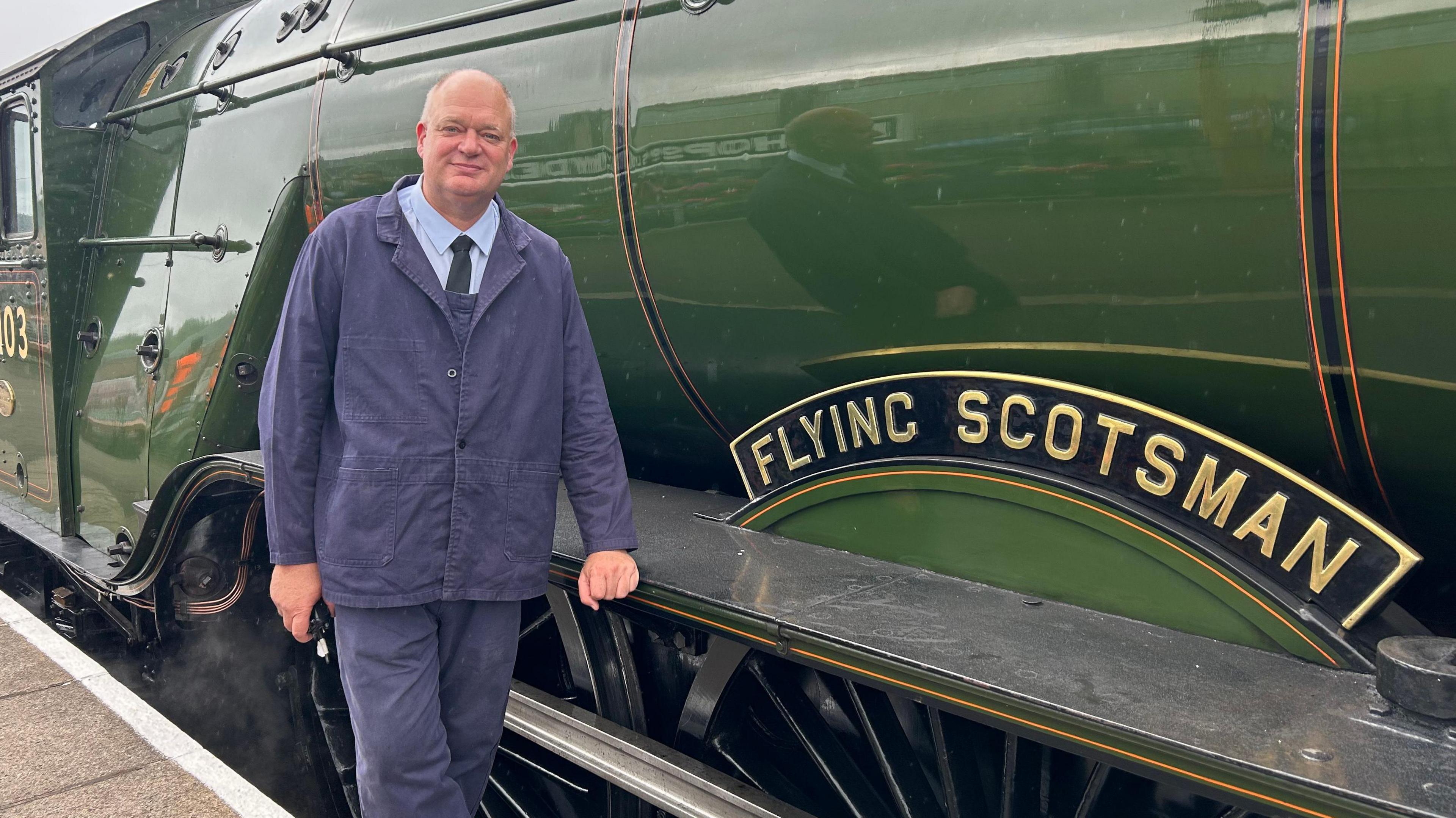 A man in a blue suit is standing next to a locomotive which says Flying Scotsman on it. It is dark green.