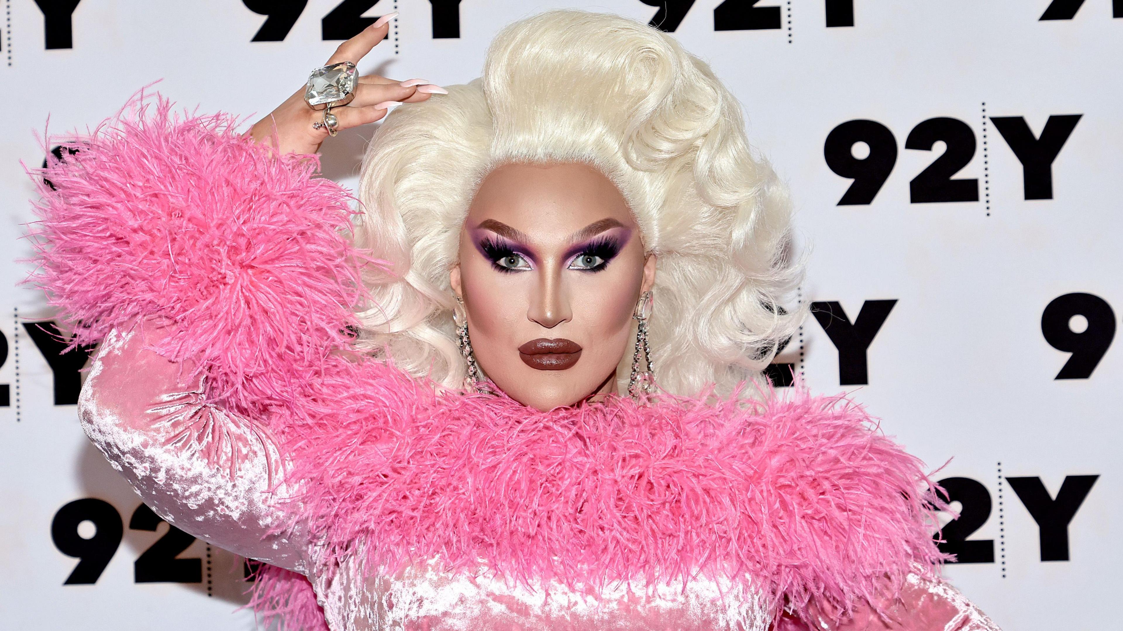 The Vivienne pictured at an event in New York in 2022. She is in full drag, wearing a pink frilly top, and looking straight at the camera with her hand above her head