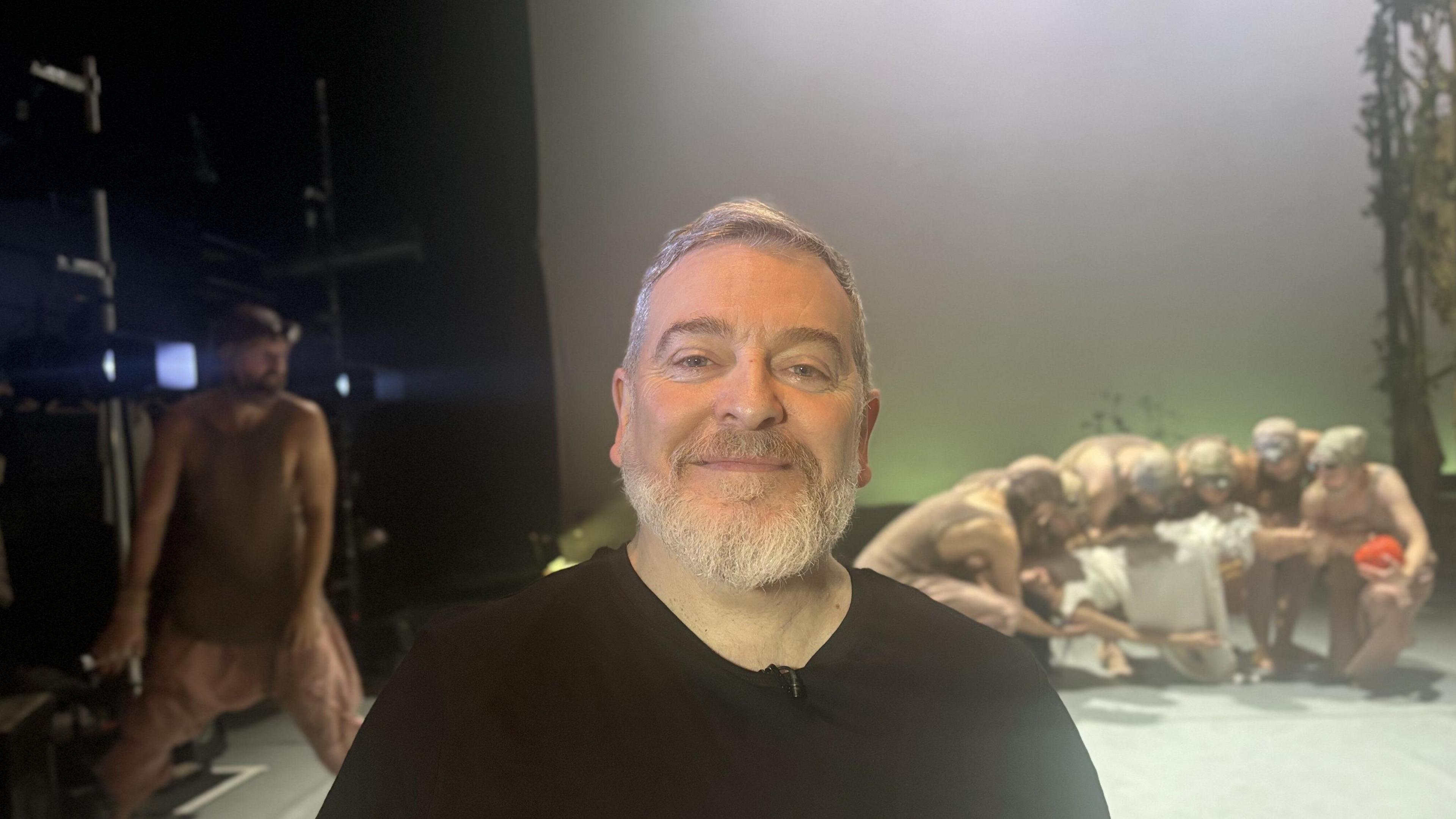 Geoff Hopson smiling in the foreground wearing a black jumper while rehearsals for Snow White: The Sacrifice go on behind him at the Northern Stage in Newcastle