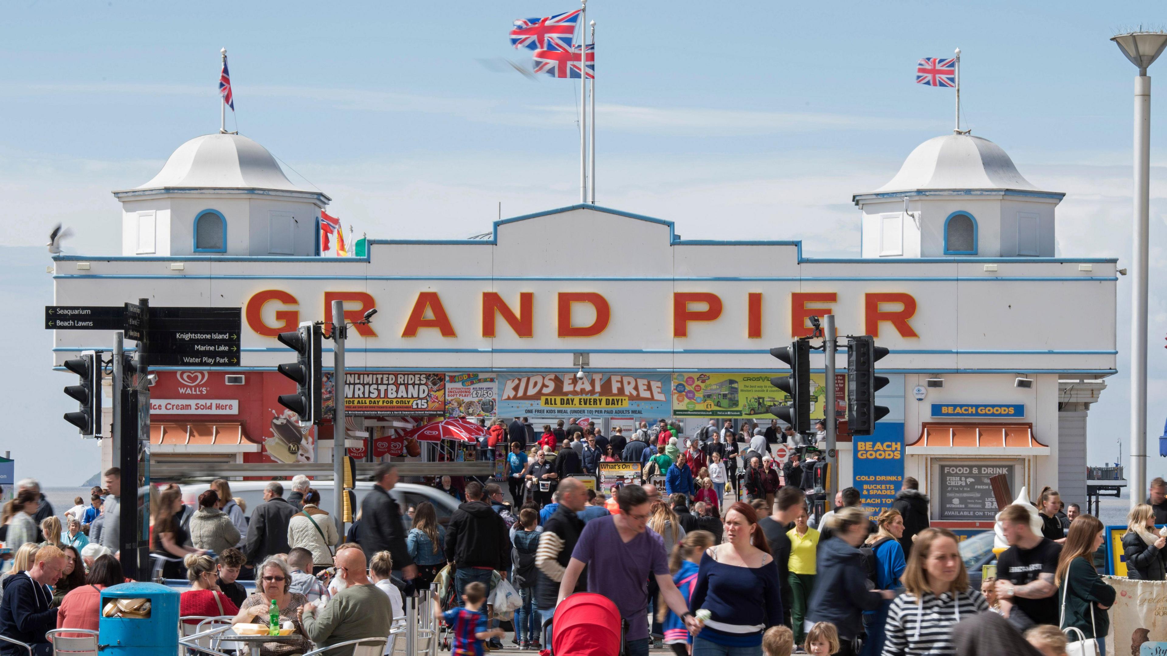 The Grand Pier in Weston-super-Mare. The pier has a white arch and union jack flags on top of it. "Grand pier" is written across the top in red capital letters and there are various signs advertising food and drink stalls. There is a large crowd in front of the pier.