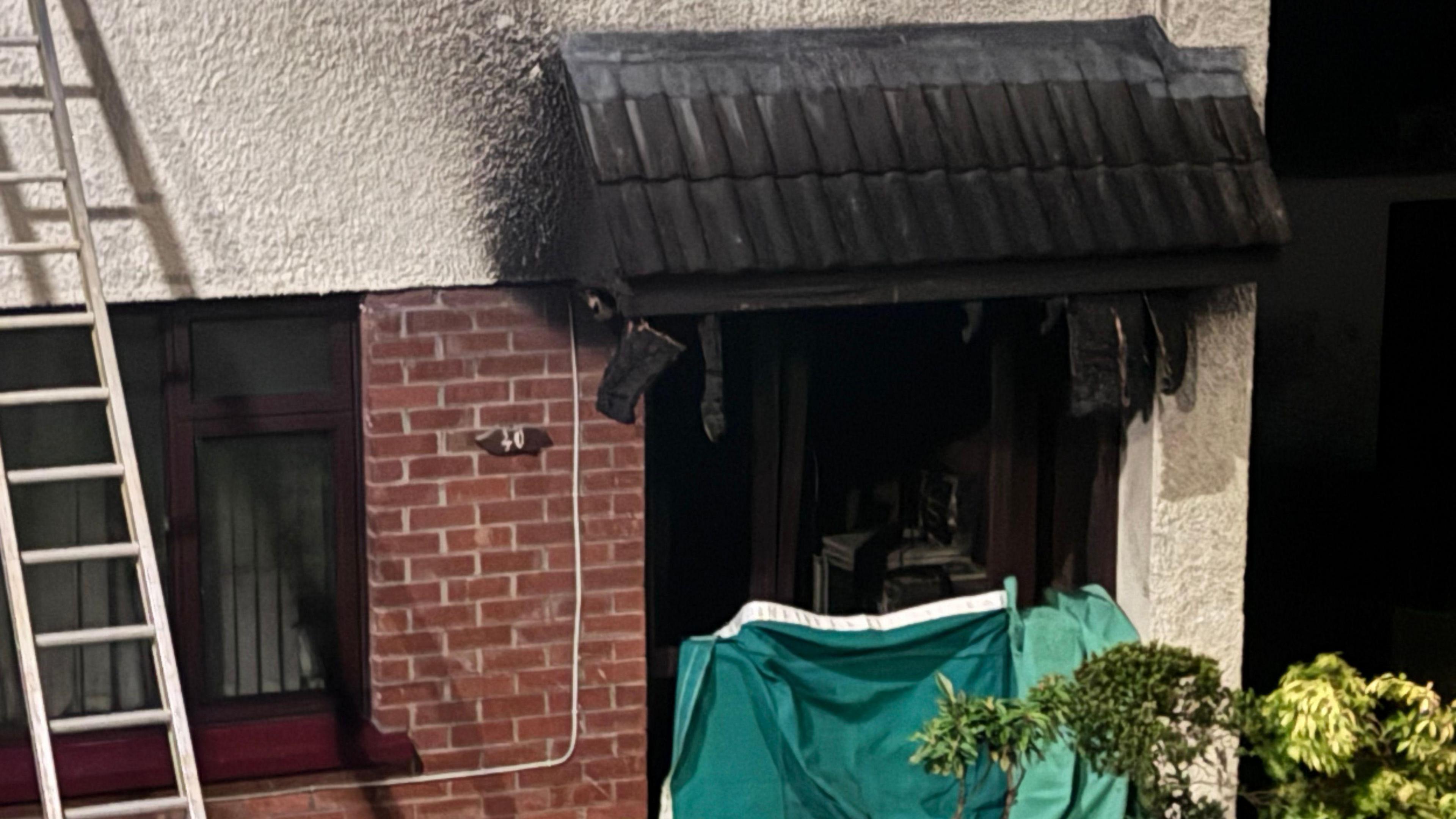 A semi-detached house with white walls and a ladder balancing against the brick wall of the window. The frame of a front door is black with ash and there is a green cover where the door used to be.