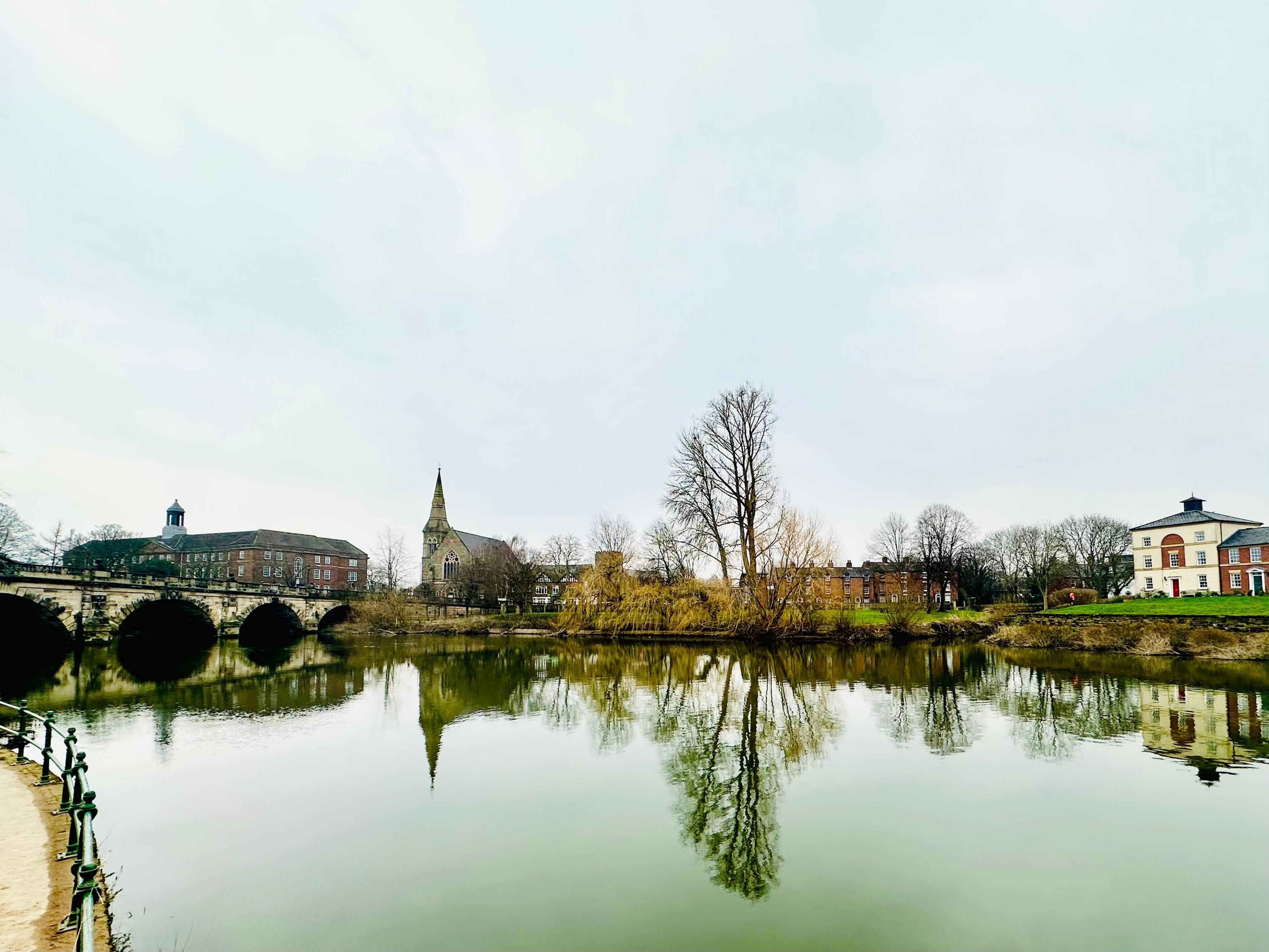 Four arches of a stone bridge span the river, with a 1930s college building, converted church and two stretches of housing making up the backdrop beyond.  They sit beyond a stark light grey sky, with the whole scene reflected perfectly in the River Severn. 