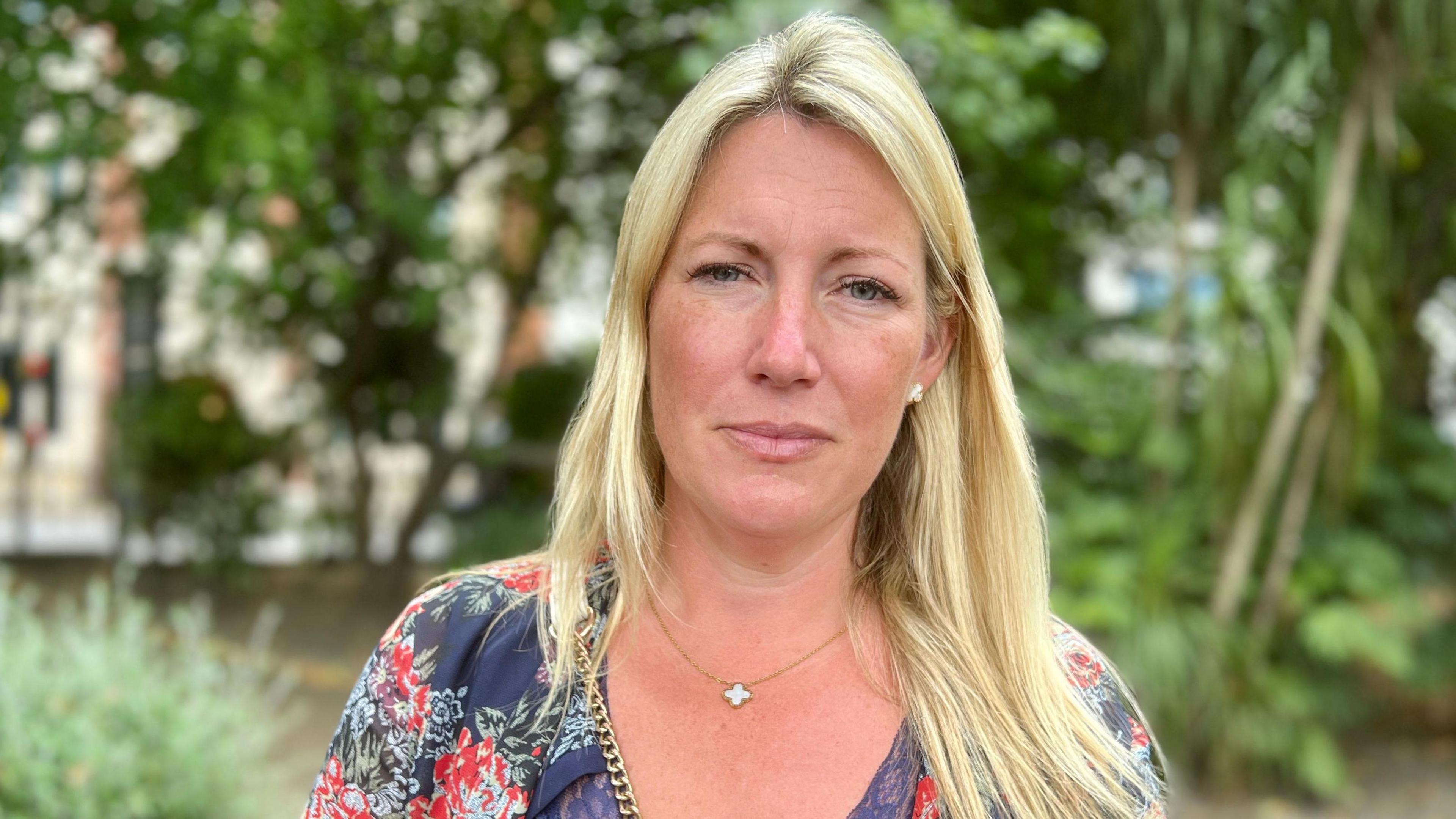 Emma Cracknell, daughter of Susan Spring standing in the gardens opposite Arundel House in central london. She is looking directly at the camera and has long blonde hair. She is wearing a navy floral v-neck top with a small cross on her necklace.