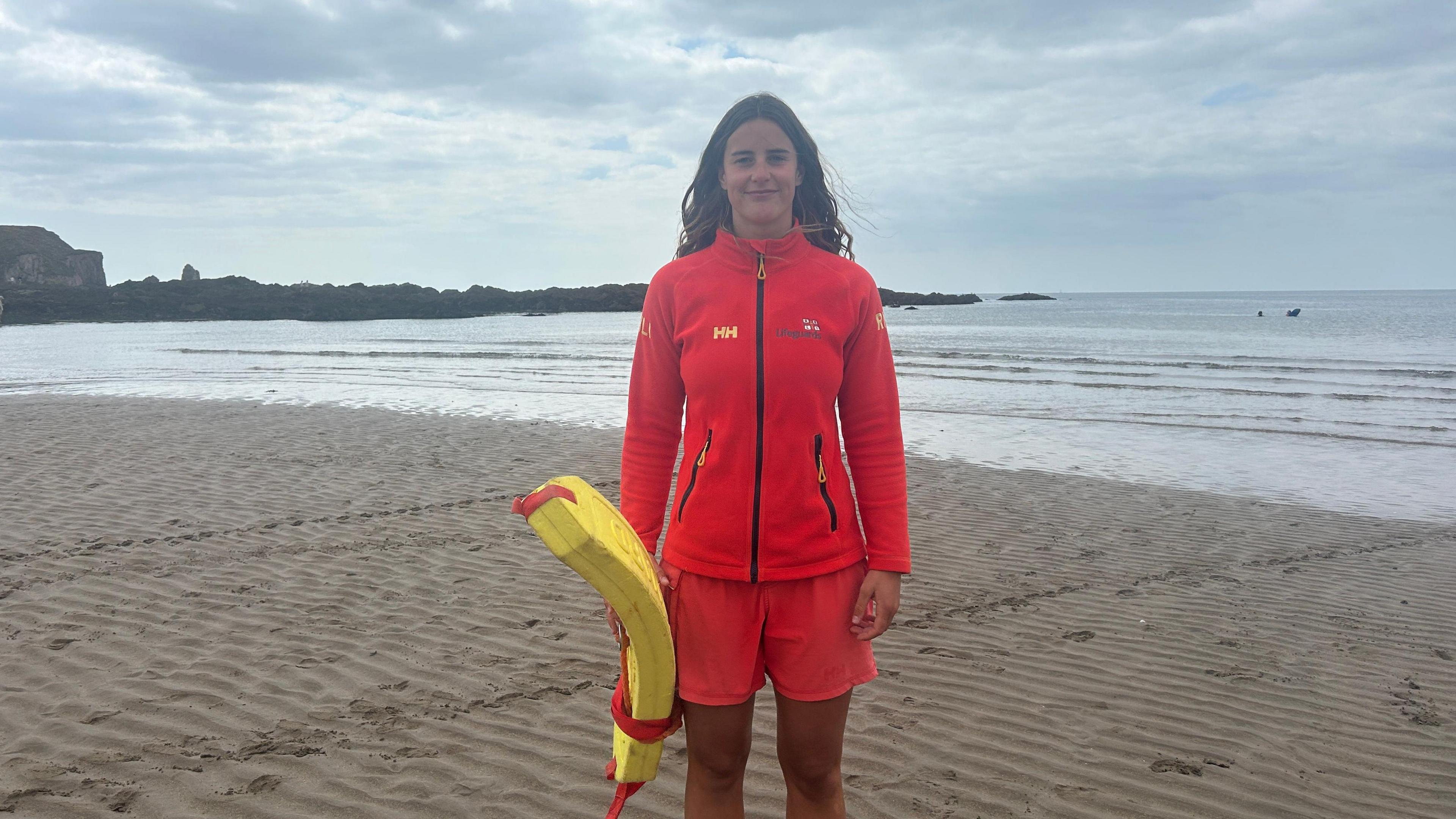 A young female lifeguard wearing a red fleece jacket and red shorts, standing on the beach. The sea is rippling onto the sand behind her. 