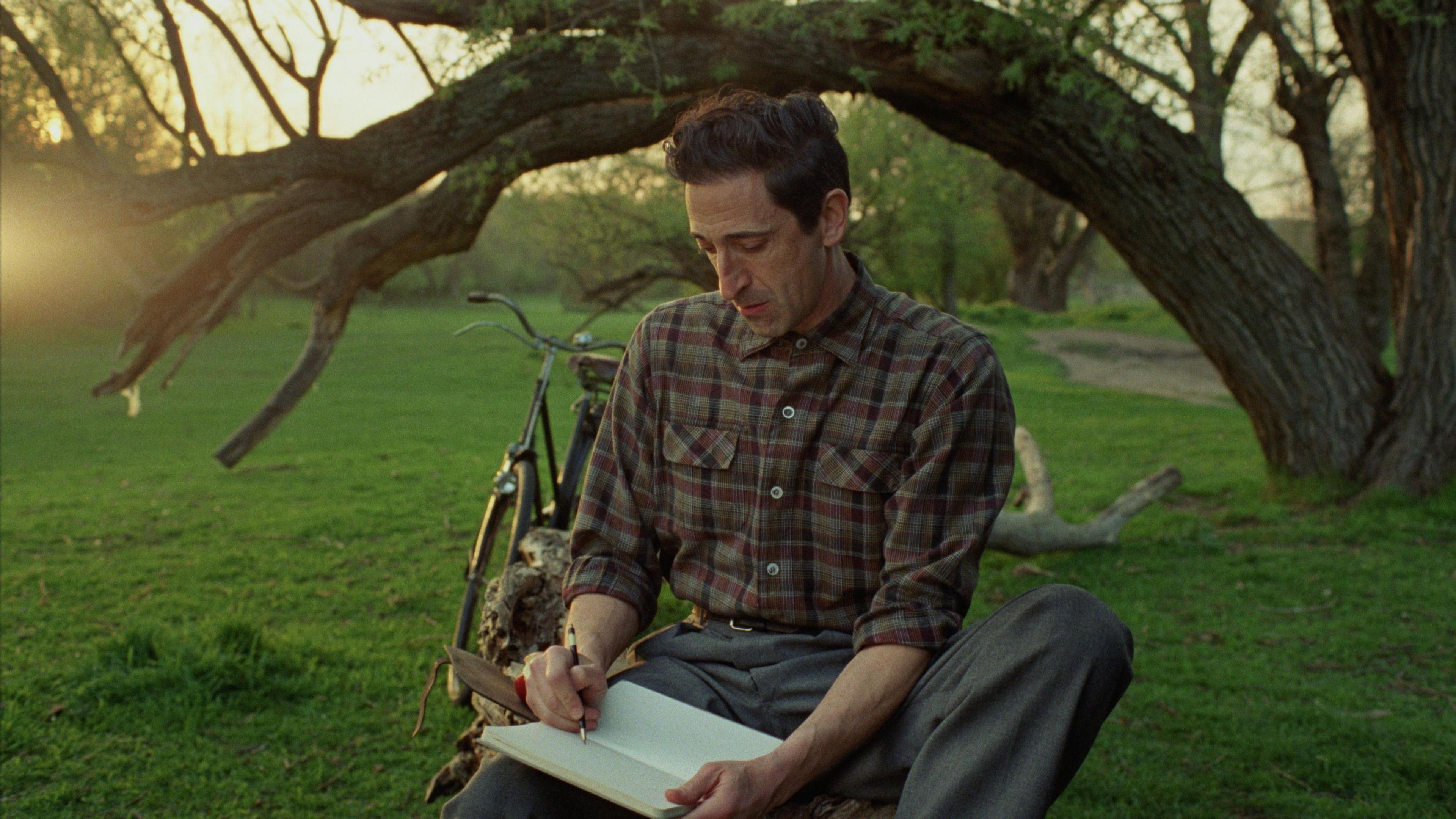 Adrien Brody sat under a tree in a grassy area. He is wearing a check shirt and grey trousers and is looking down at a notebook, which he is writing in.