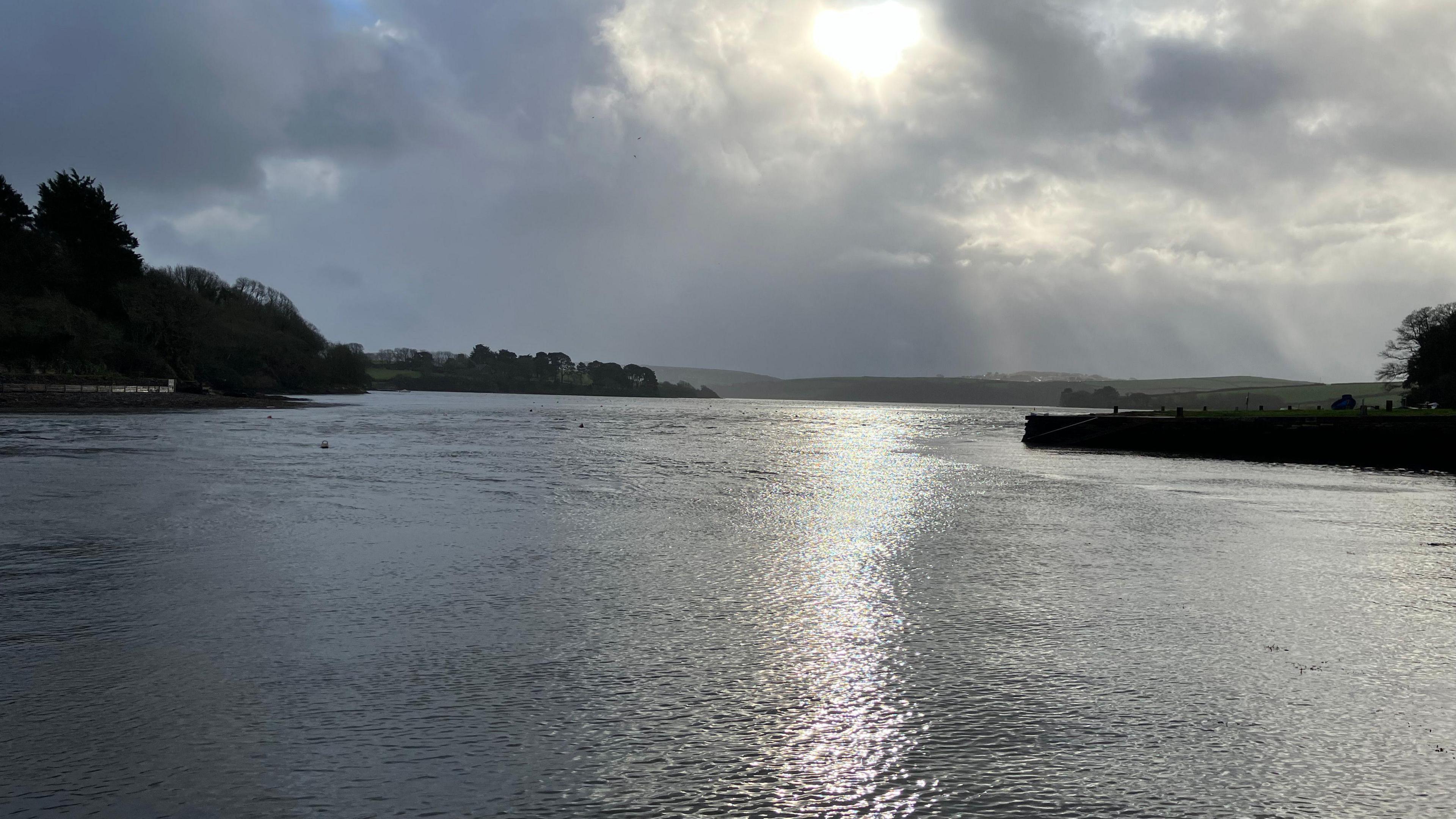 Kingsbridge estuary with the sun setting. It is a cloudy day and in the distance there is land either side of the estuary. The sun is coming through the clouds at the centre and is reflecting off the water.