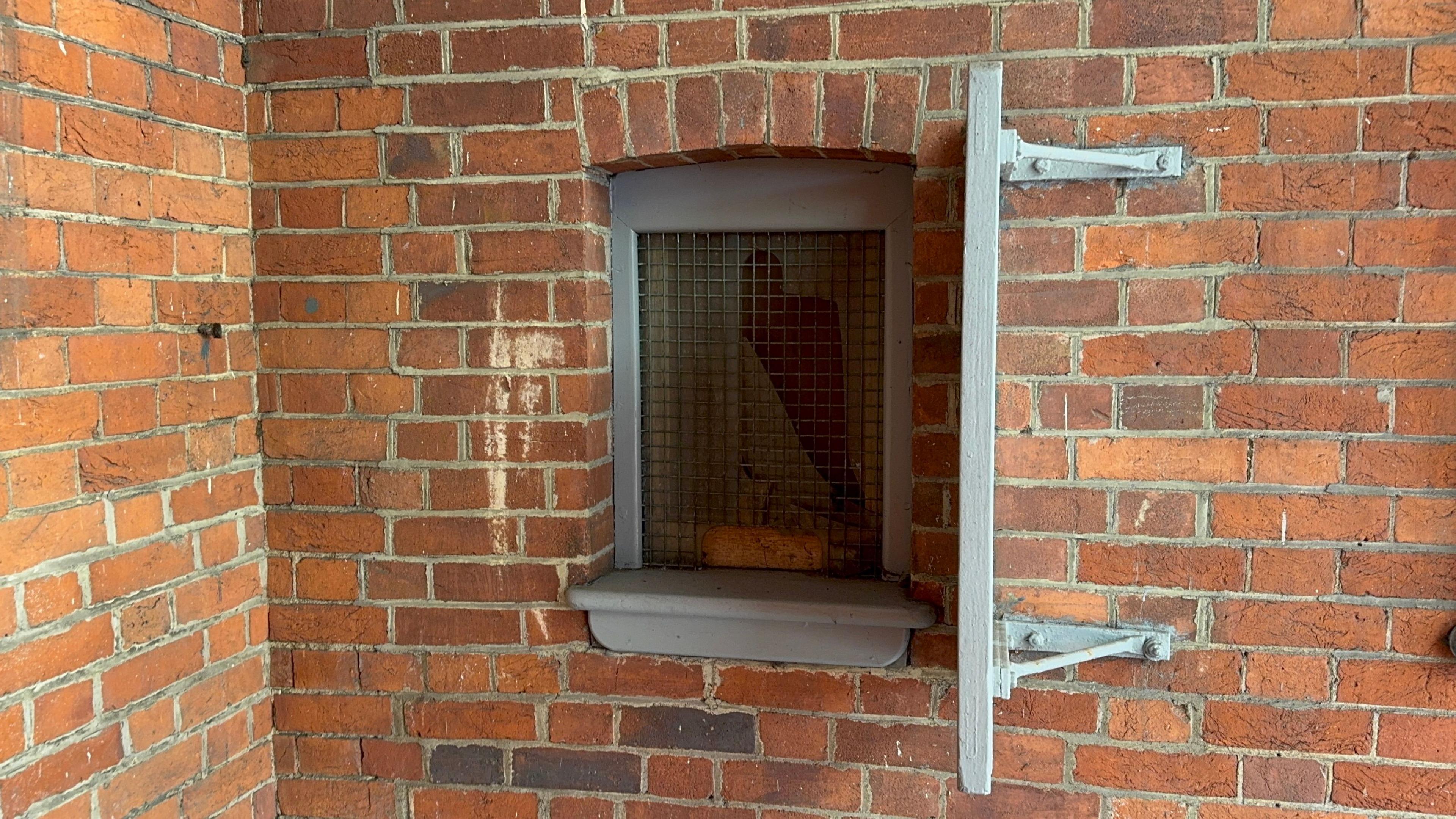 A grey slot window in the middle of a brown brick wall. It is covered in grey metal mesh with a thin windowsill. 