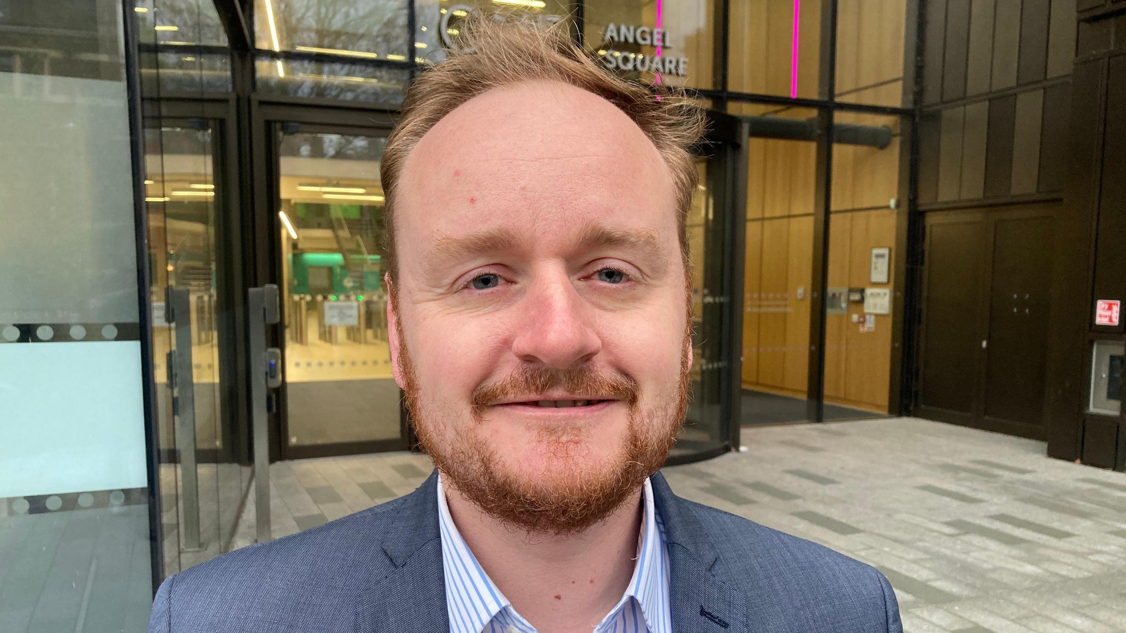 Adam Brown wearing a blue suit with a white and blue pinstriped shirt. He is standing in front of One Angel Square - the headquarters of West Northamptonshire Council.