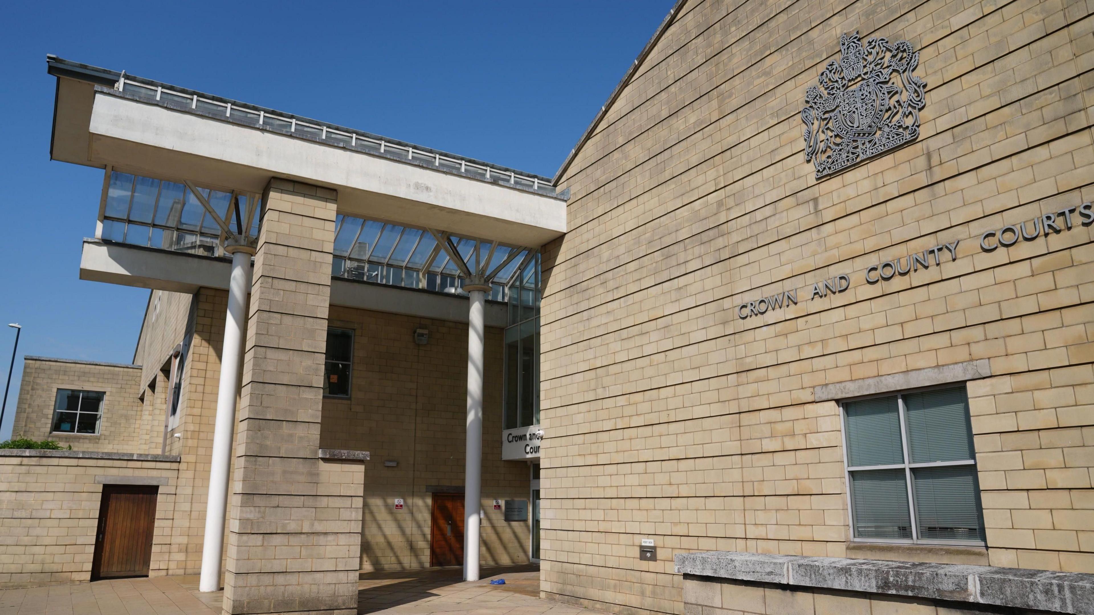 A cream brick building with the silver crest of a Crown Court on it, pictured on a sunny day.