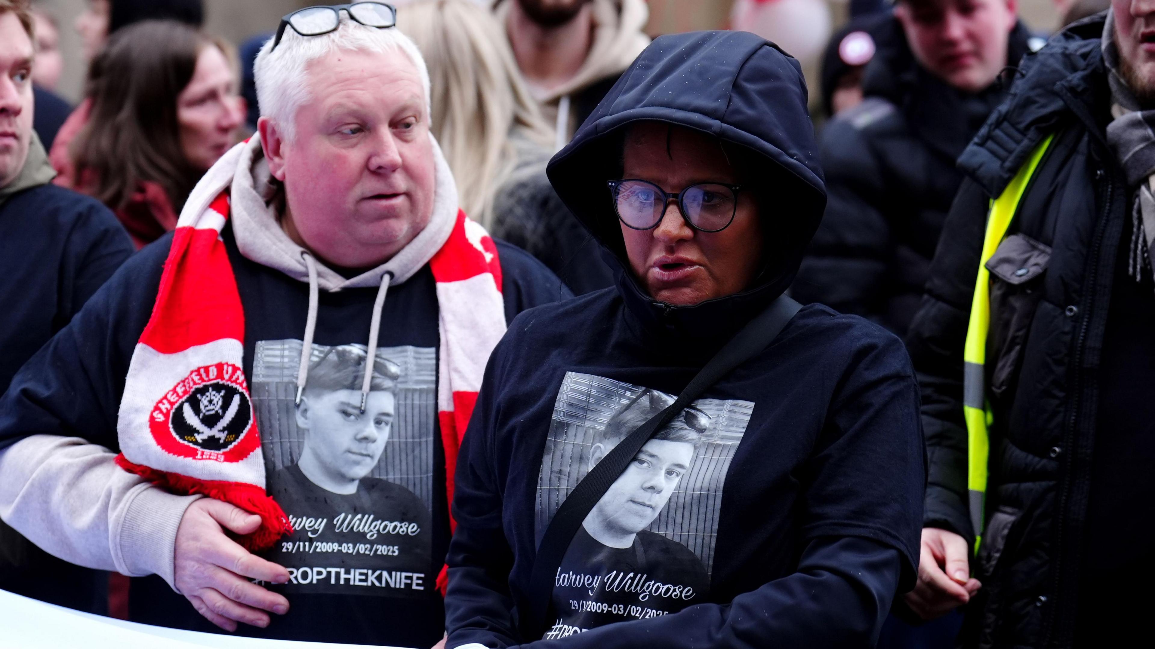 Mark Willgoose and Caroline Willgoose wearing t shirts with their son's face and the slogan 'drop the knife' on it. Mark is also wearing a Sheffield United scarf.