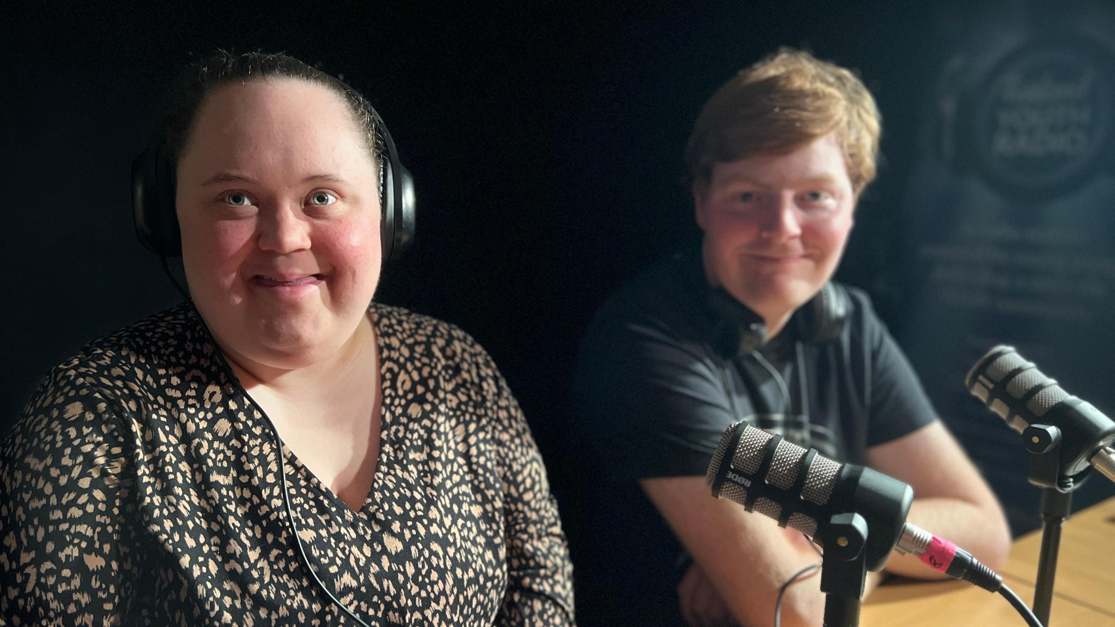 Two podcasters, a young woman and a young man, sit at a desk facing the camera with a microphone in front of each of them.