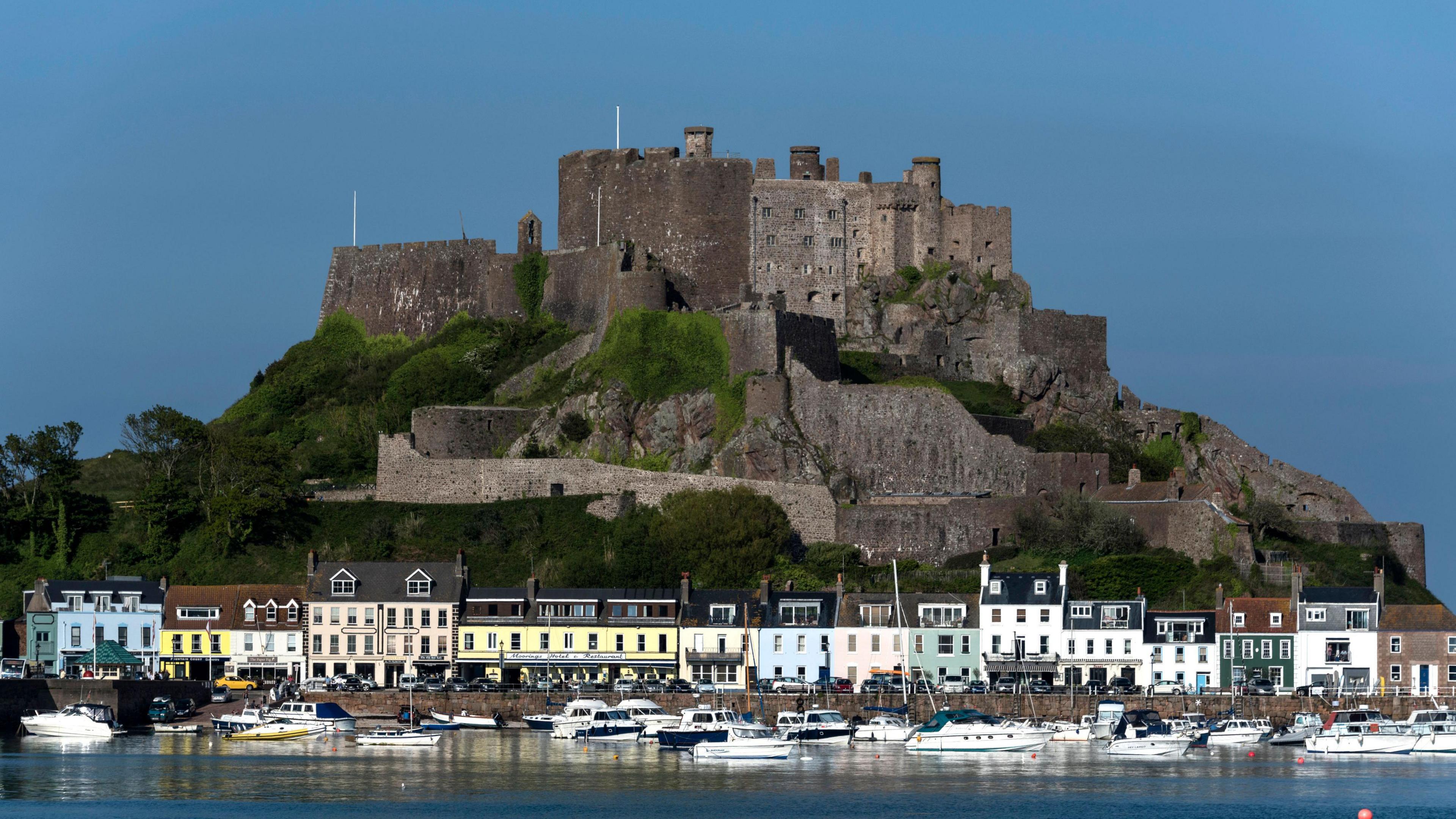 A castle on top of a hill overlooking a harbour full of boats, mainly white speed boats, and a row of terrace buildings.