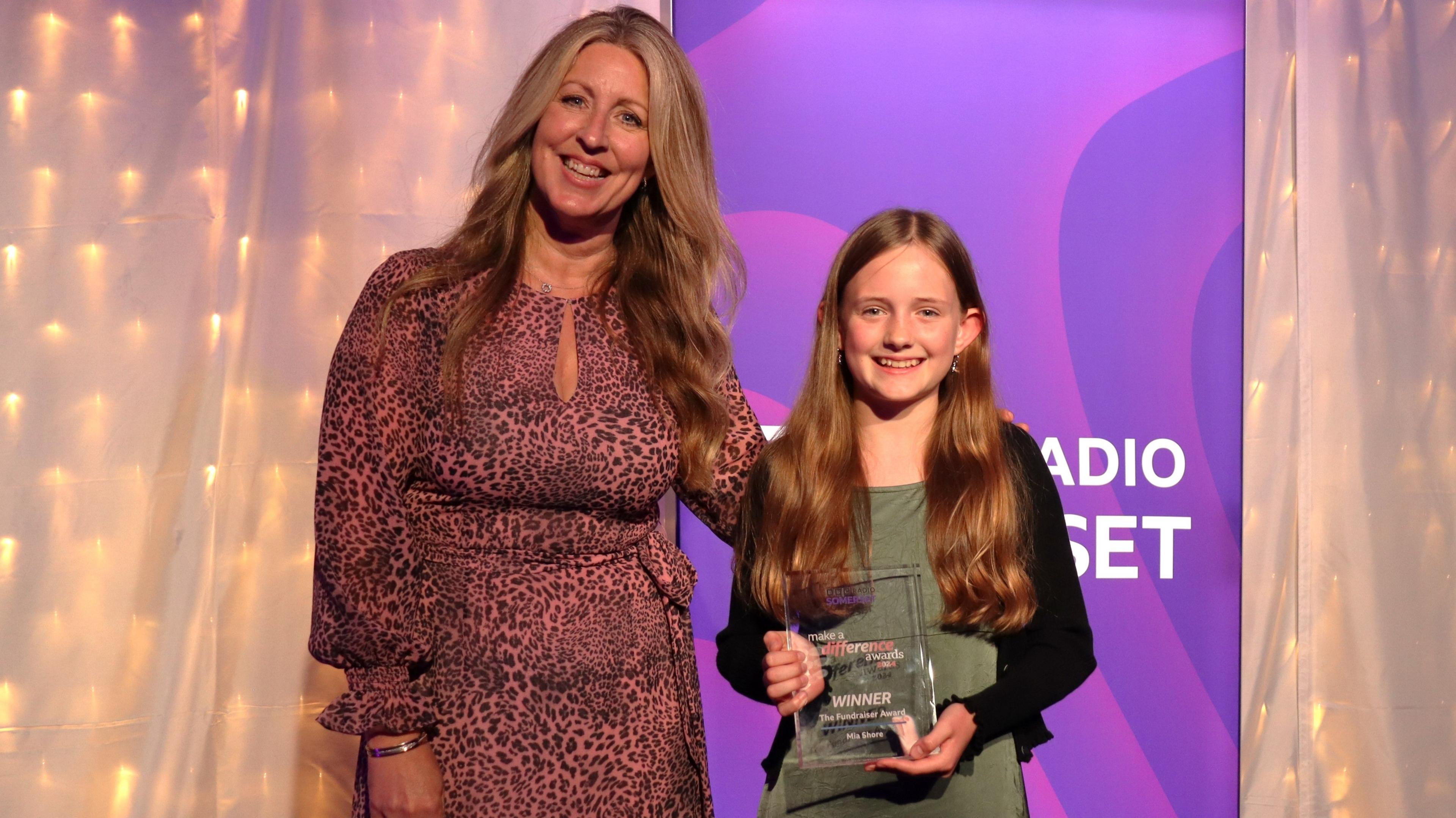 Mel Everett wearing a pink leopard patterned dress, standing beside Mia with her arm around her. Mia is wearing a green dress and a black cardigan, holding her award and smiling. In the background there is a pink BBC Radio Somerset banner and a white sheet with fairy lights underneath it.