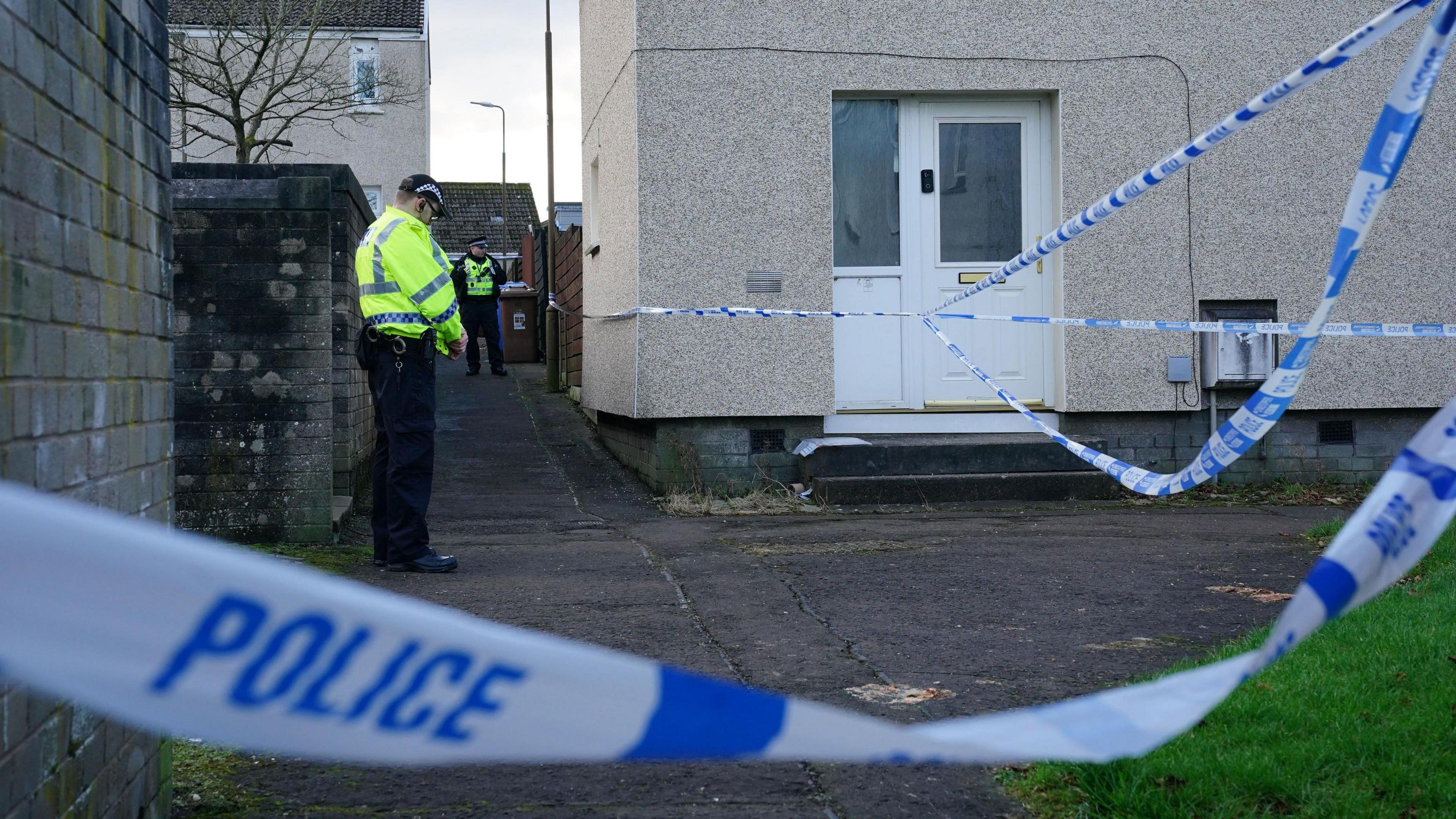 A police cordon at the scene in Harburn Drive, West Calder. An officer wearing a hi-viz jacket can be seen in the garden. The house is grey with a white door. In the foreground, there is white and blue police tape.