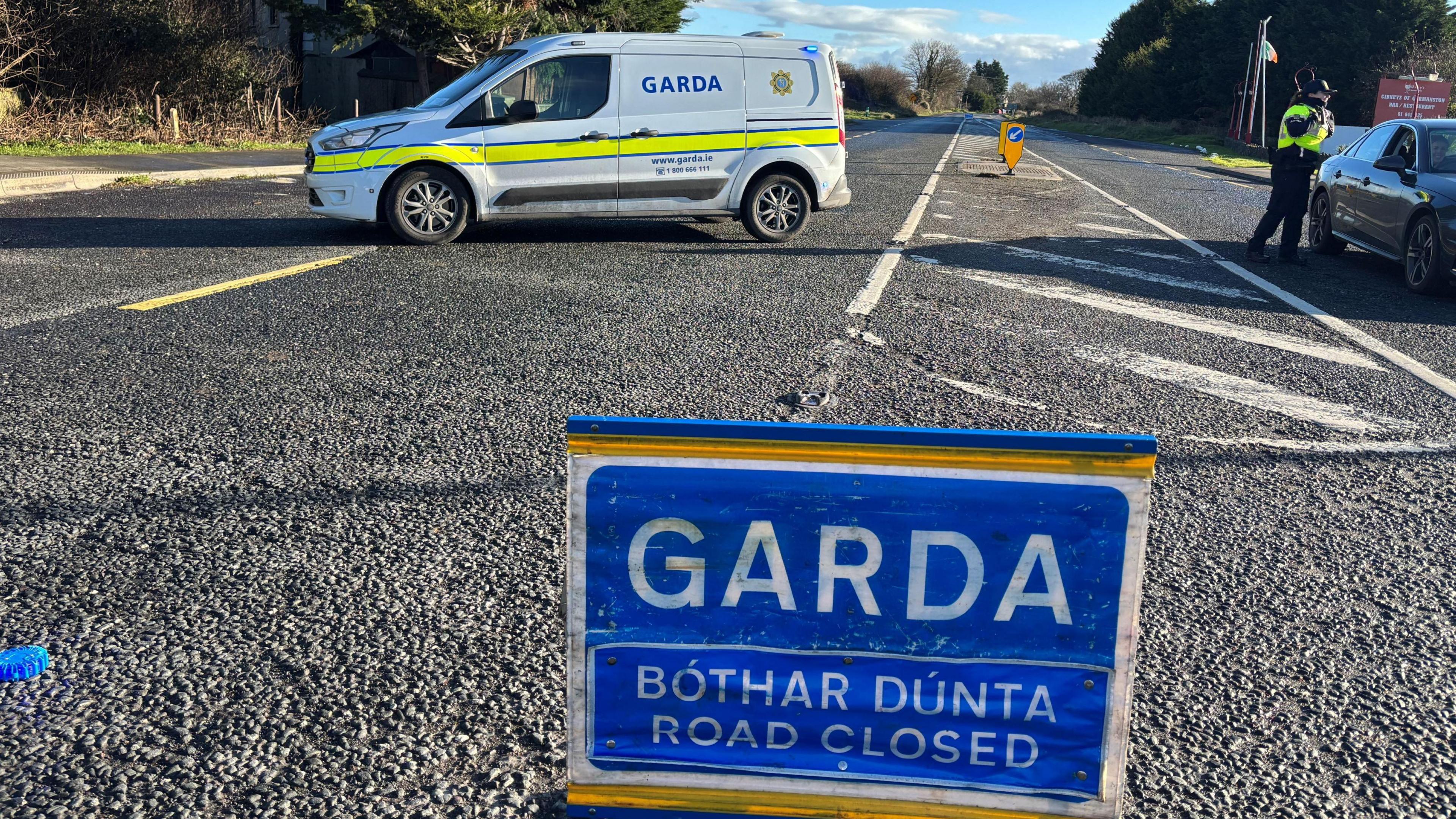 A blue Garda road closed sign sits in the middle of a road. In the background, there's a white van with Garda written on it and an officer is speaking to the driver of a grey car. It is a bright day with blue sky off in the distance.