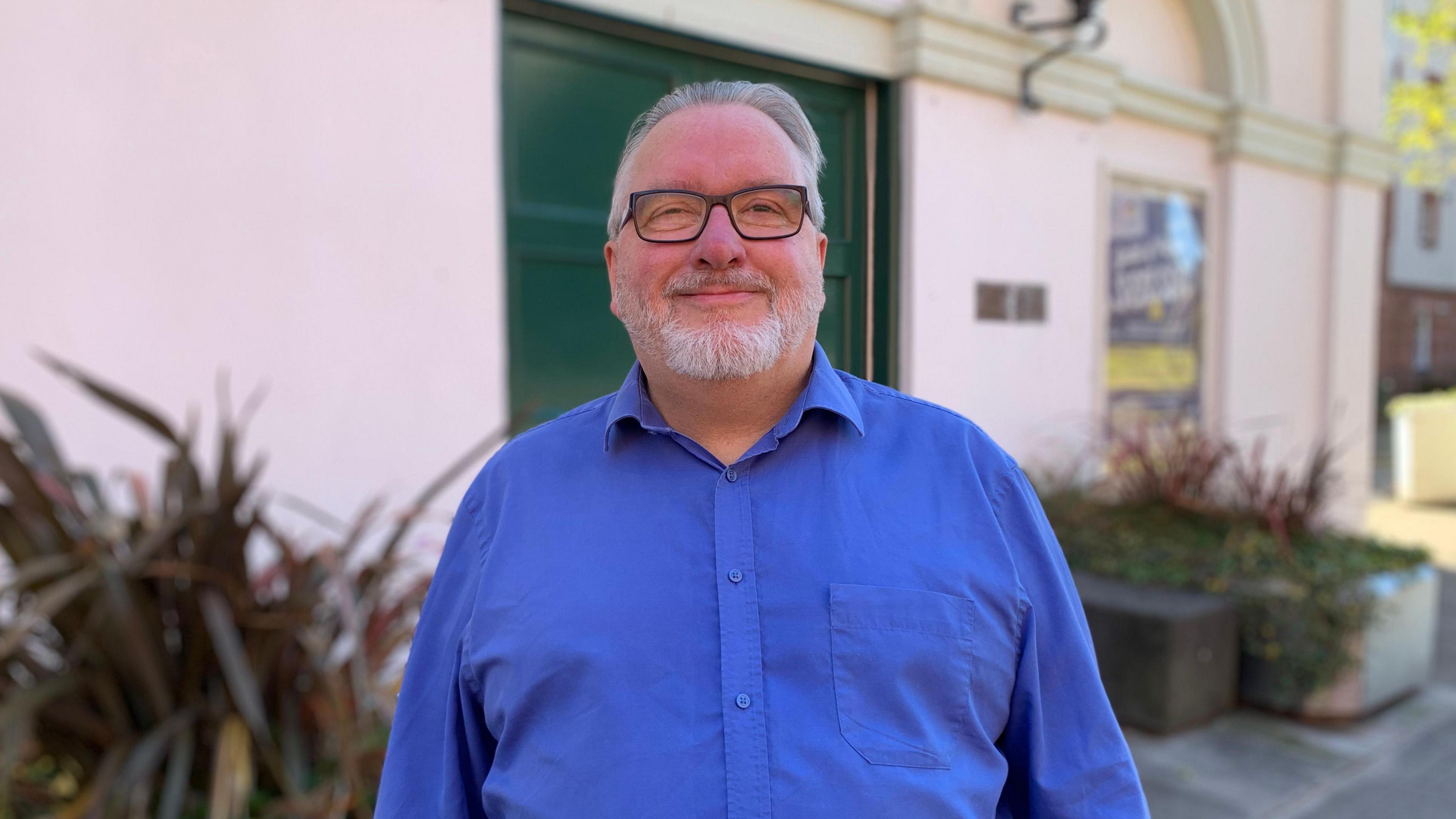 A man in glasses with grey hair and beard in a blue shirt stands in front of a theatre building