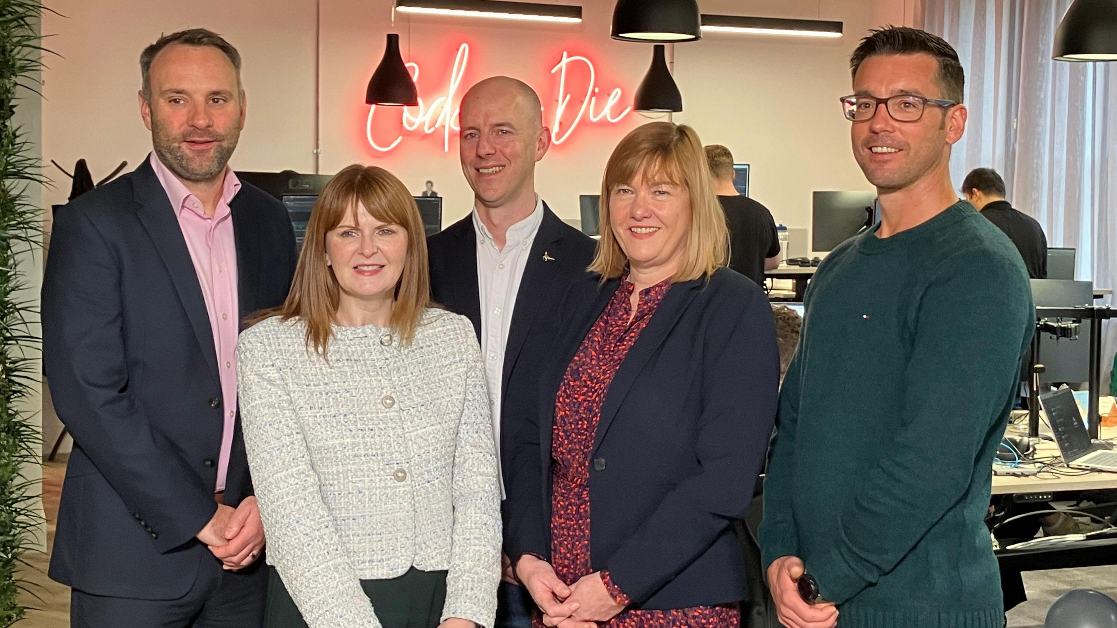 Caoimhe Archibald stands as part of a group of two women and three men in an office space. Each of them smile towards the camera and a red neon sign hangs of a wall in the background.  