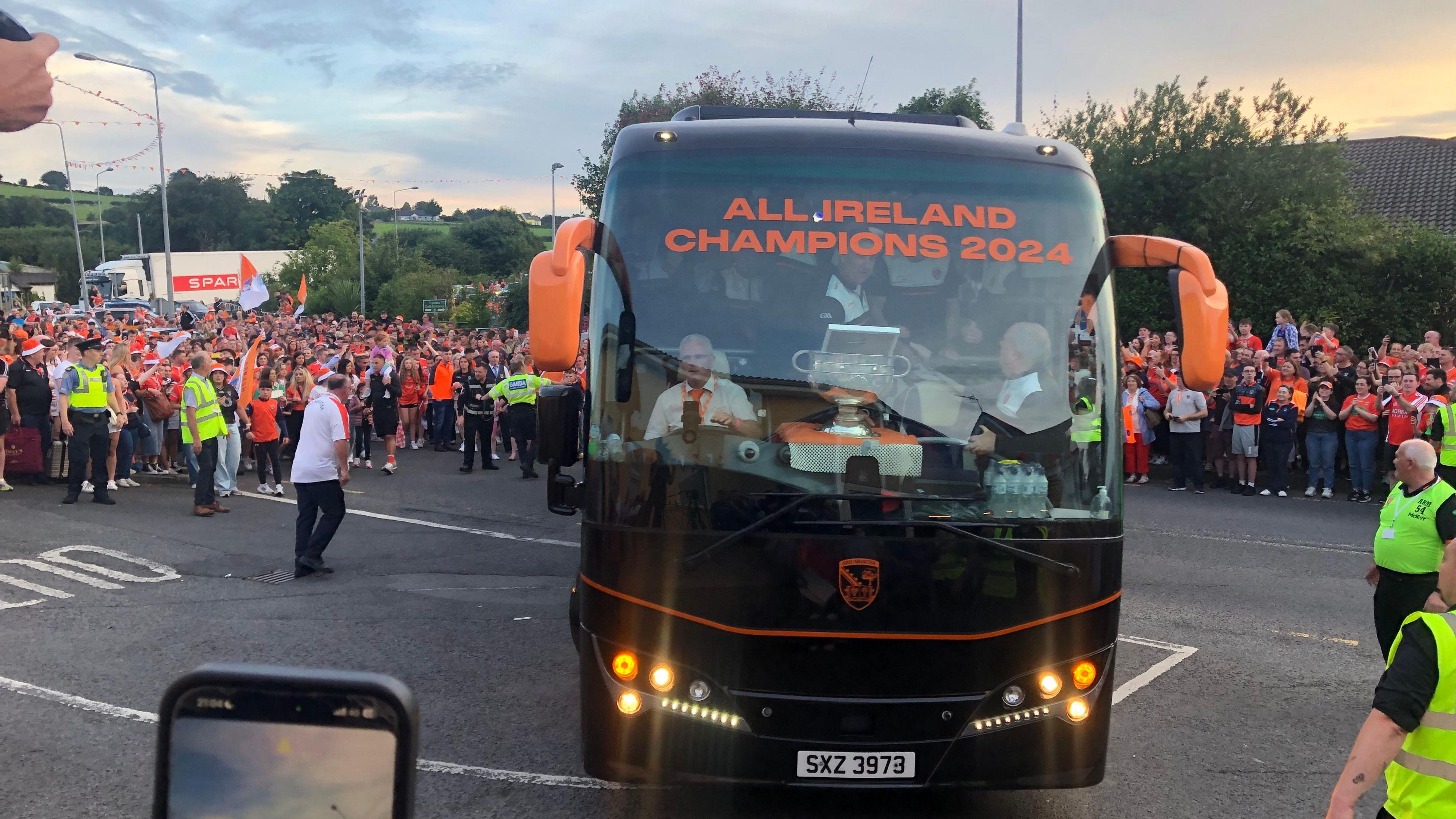 Bus with the words "All Ireland Champions 2024" with the Sam Maguire cup in the front window. A  large crowd of people in the background.