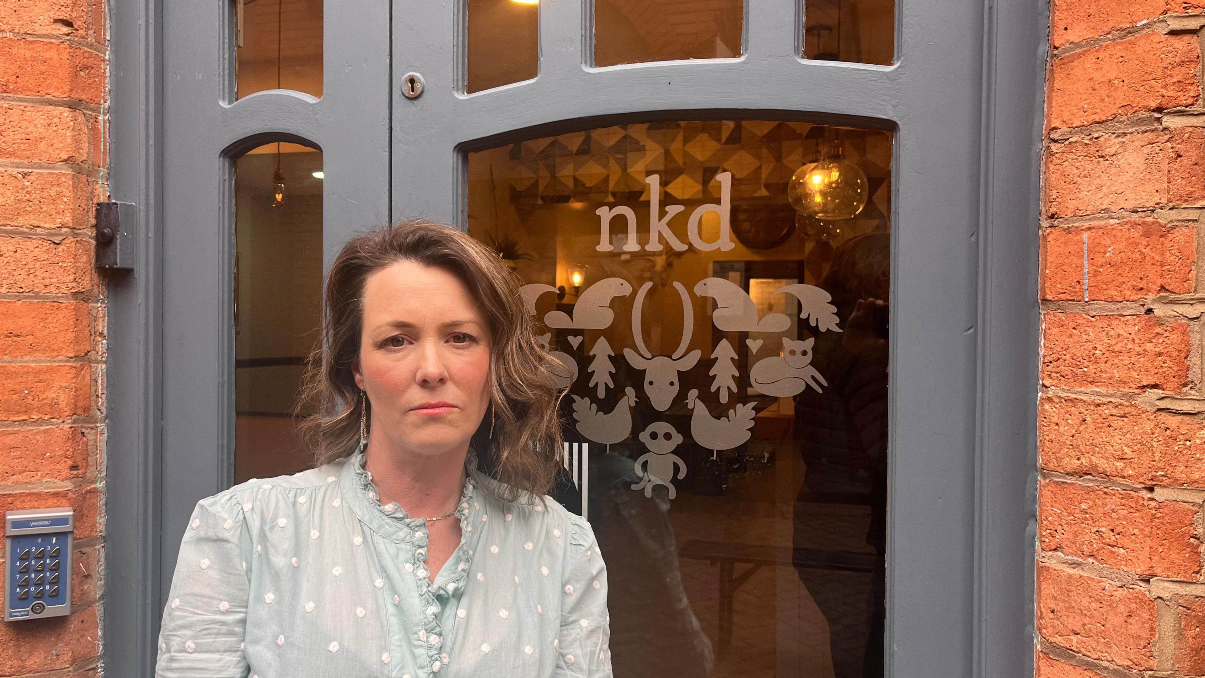 A woman with long dark hair, wearing a blue shirt with spots, standing in front of a salon