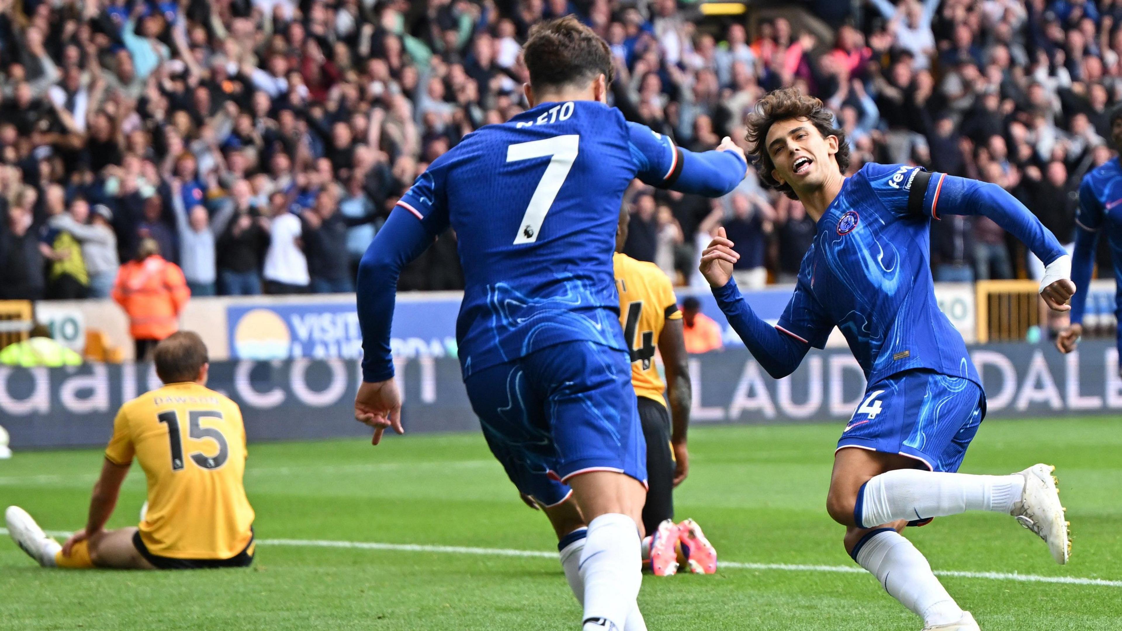 Joao Felix, on the right of the picture, turns to face Pedro Neto while running with Neto in the foreground of the shot with his back to the camera after setting up Felix for Chelsea's sixth goal. Craig Dawson sits on the left of the shot with his back to the camera and hands on his knees in disappointment. In the background Chelsea fans celebrate at Molineux.