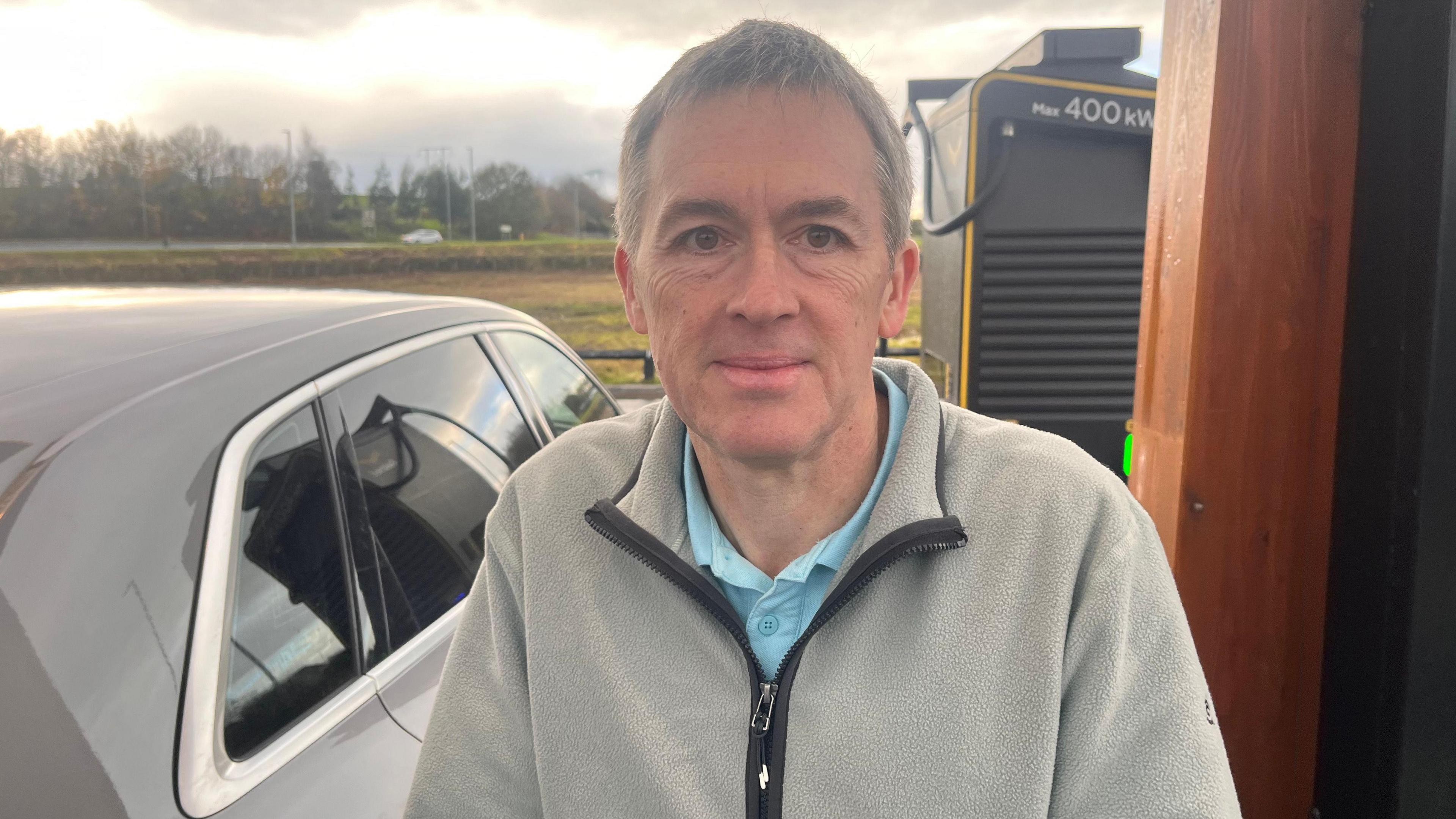 Jason is standing next to his car. He's wearing a grey fleece.