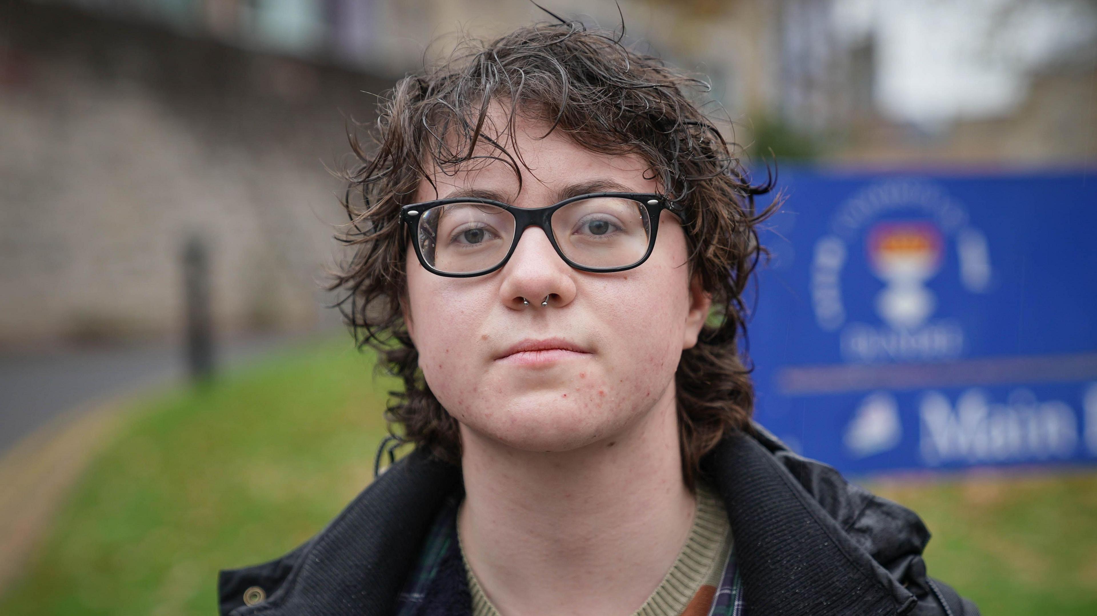 Michael McCarey, who has short, dark, curly hair and glasses, stands outdoors, wearing a dark jacket. A blue sign with a crest and text is visible in the background.