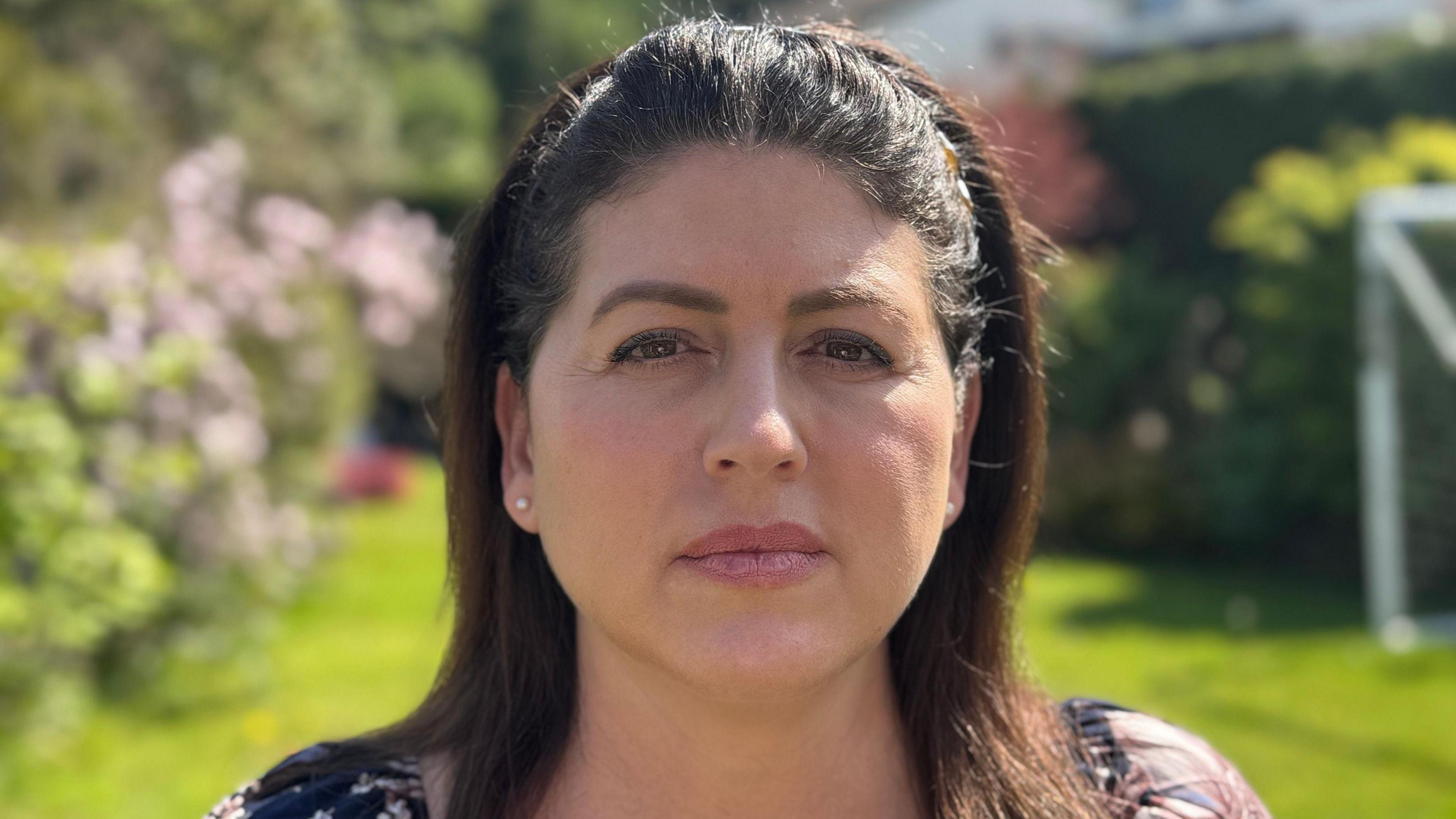 A woman stood in a garden looking at the camera. She has dark brown-shoulder-length hair.