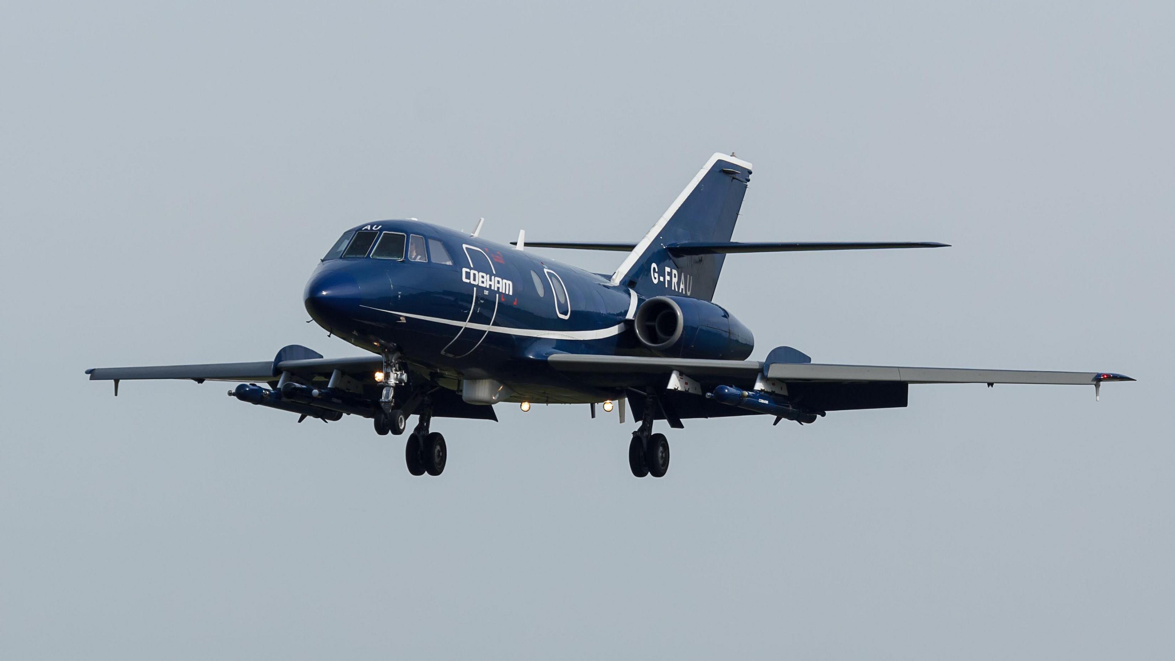 A Dassault Falcon 20 aircraft. The jet is blue with white lines. It has jet engines above its wings. Its landing gear is down.