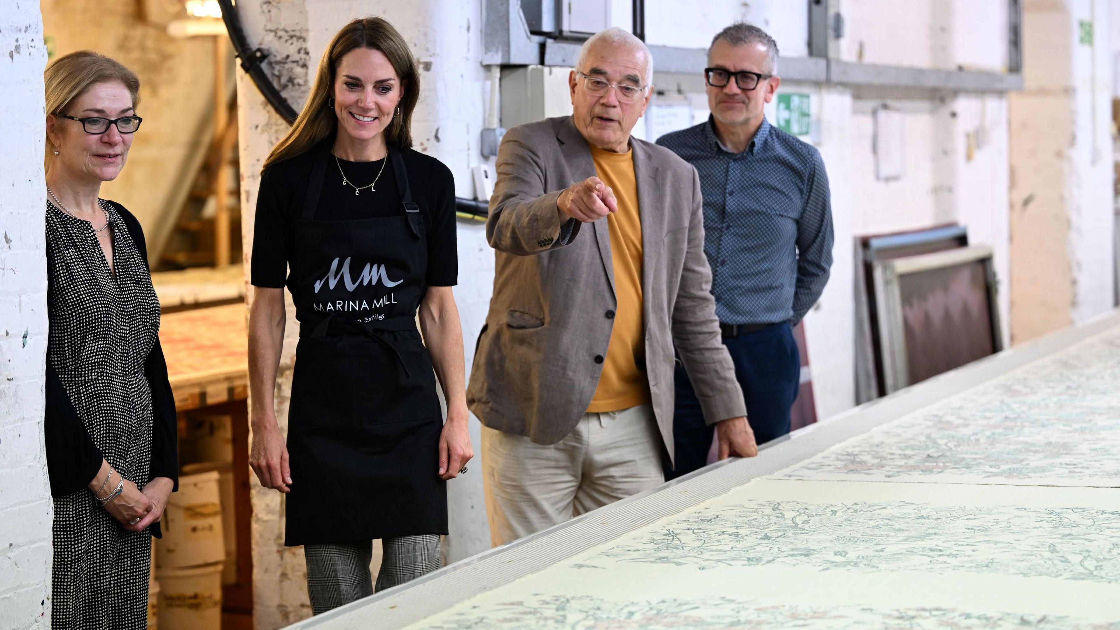 The Princess of Wales wears a black Marina Mill apron and looks toward the camera in the direction of a long printed sheet on a table. She is with three other people, two men and a woman who are explaining the item to her. 