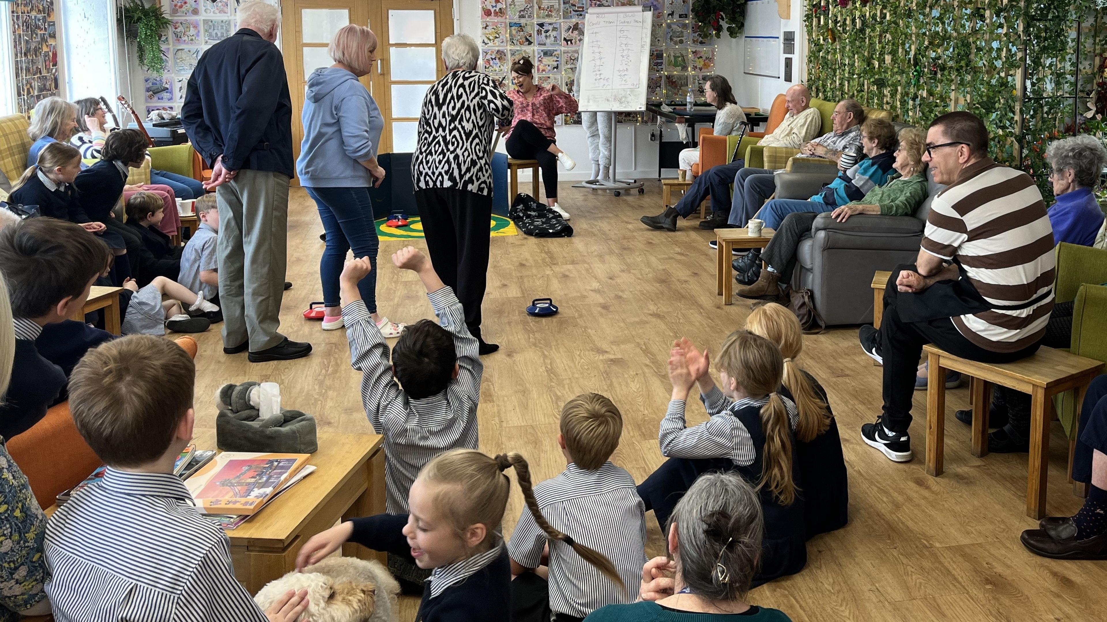 A club member pushing a curling stone towards a target with school children in the foreground celebrating and stroking a therapy dog and members of staff, club members and volunteers gathered around to watch the competition