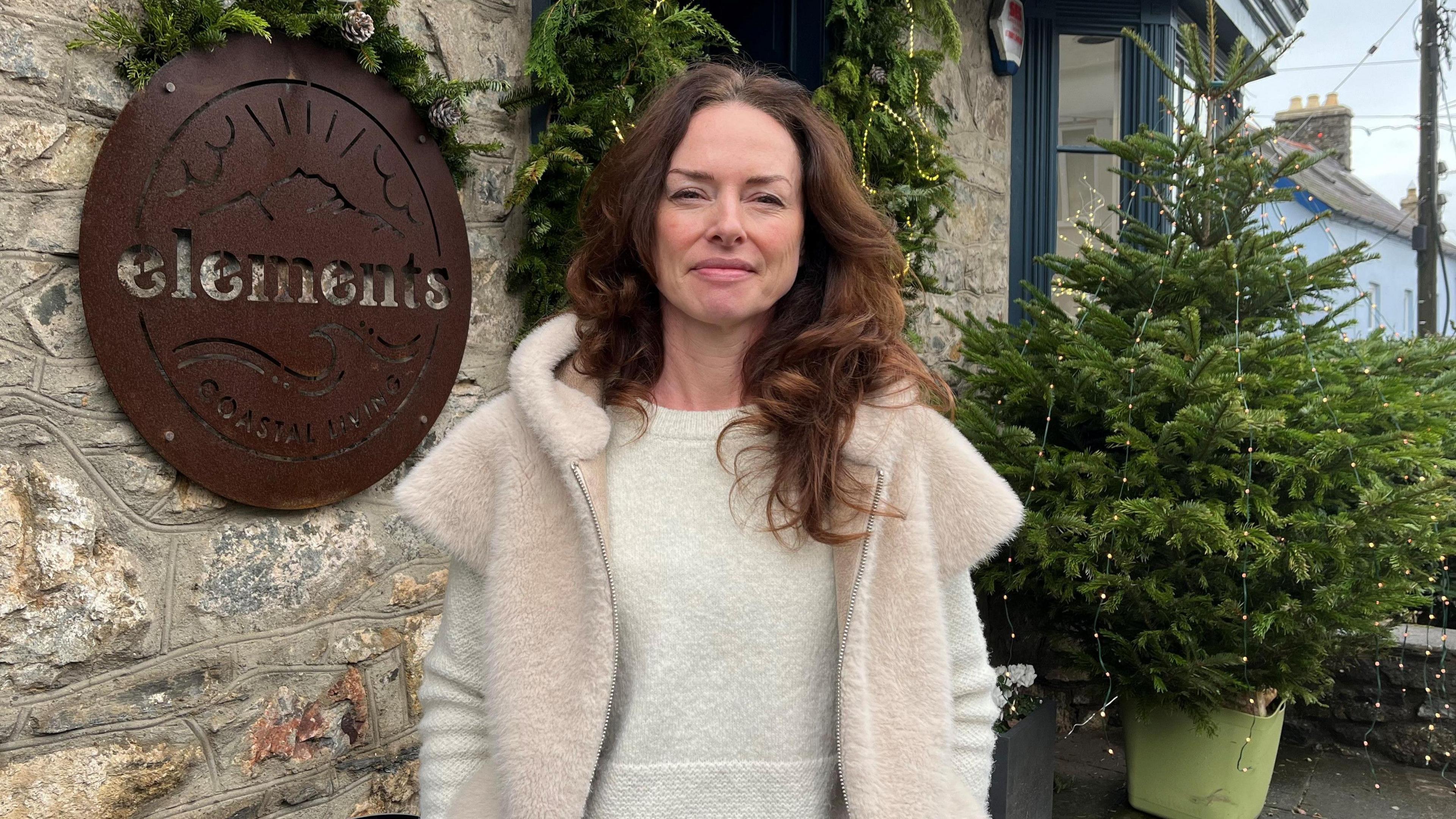 Isobel Fallows has long wavy brown hair and is wearing a pale furry fleece and white jumper. She is stood in front of a sign that says elements and a small Christmas tree