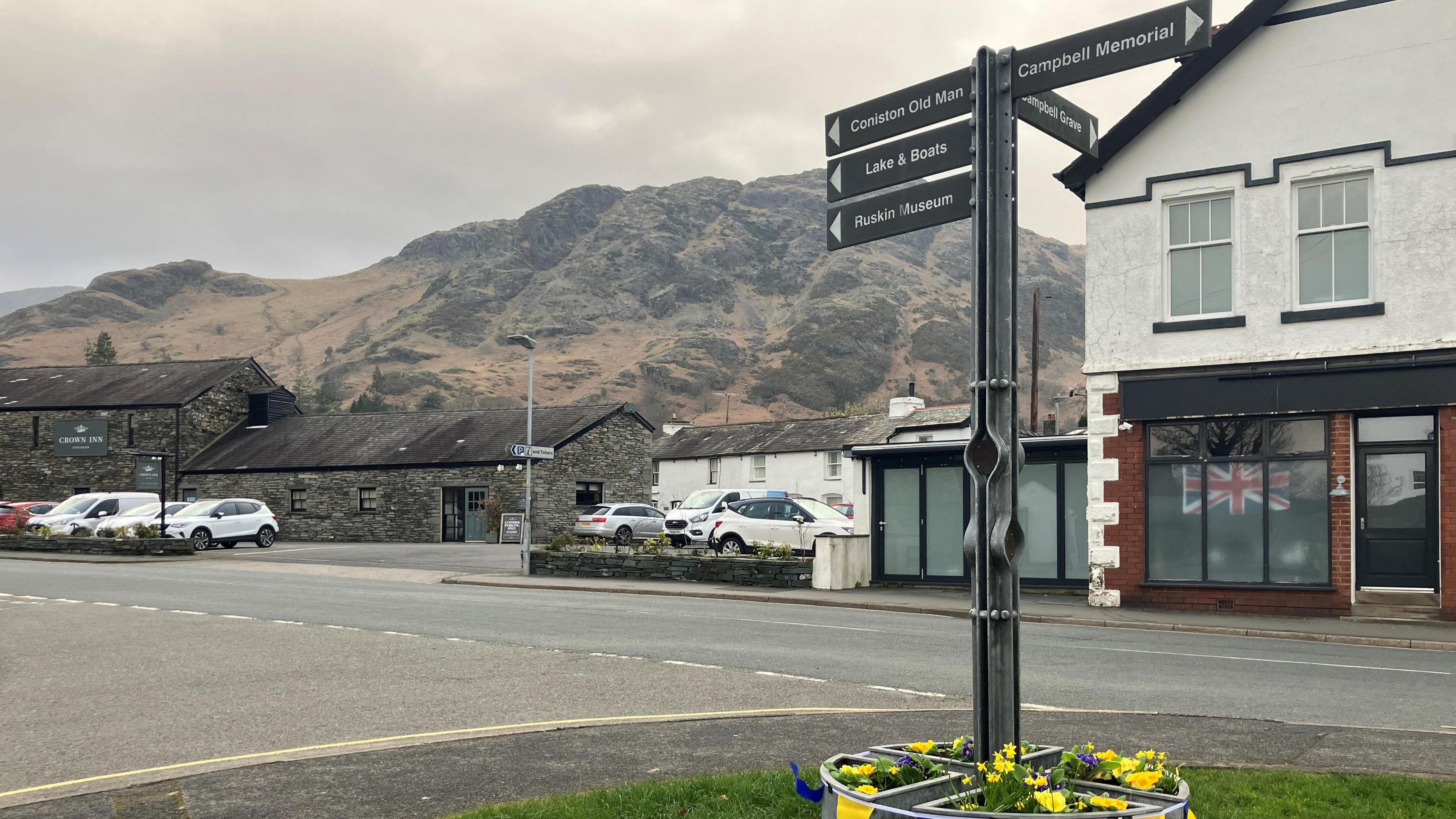 A general view of the village of Coniston. There is a sign in the foreground signposting to various attractions in the village. There are shops and buildings lining the main street and the fells in the background.