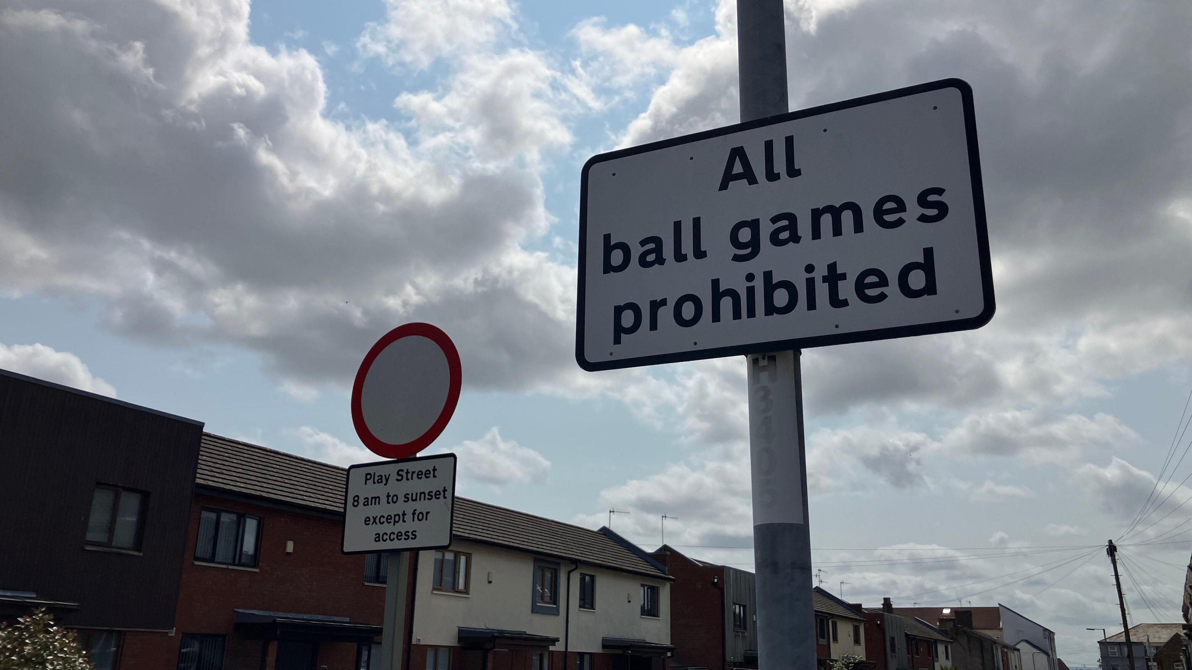 Two signs are on a residential street lined with houses. One reads 'All ball games are prohibited'. The other says 'Play Street 8am to sunset except for access'.