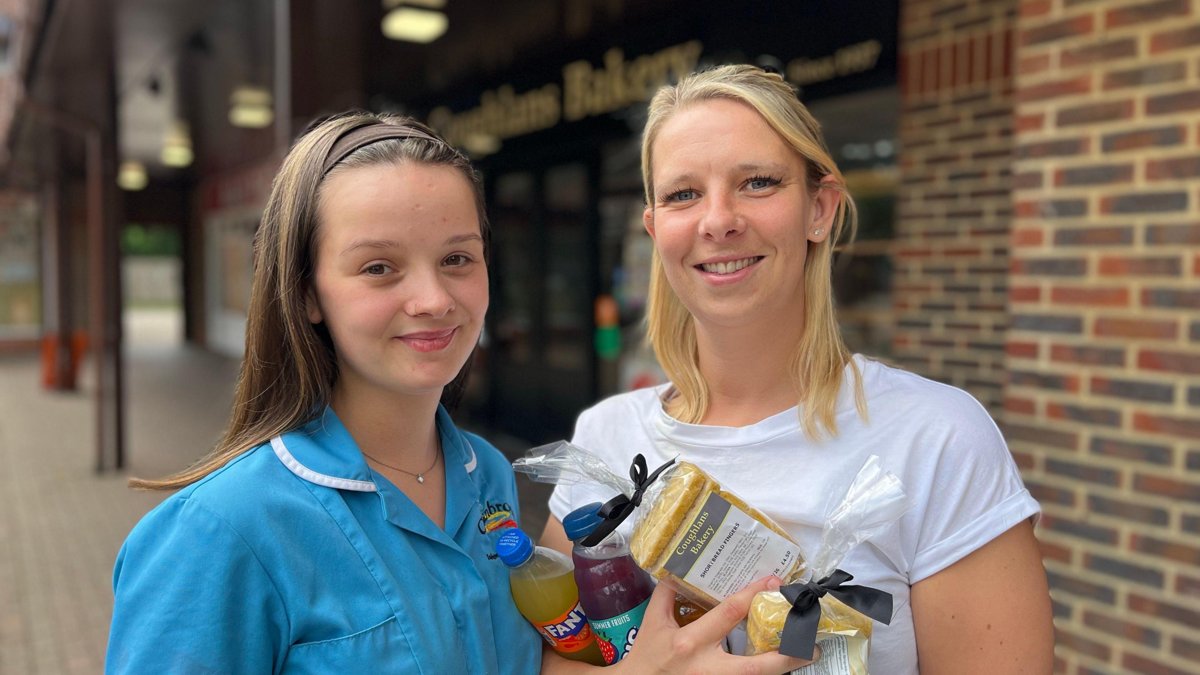 Two women smile. One is wearing bright blue overalls, the other, a white T-shirt. 
