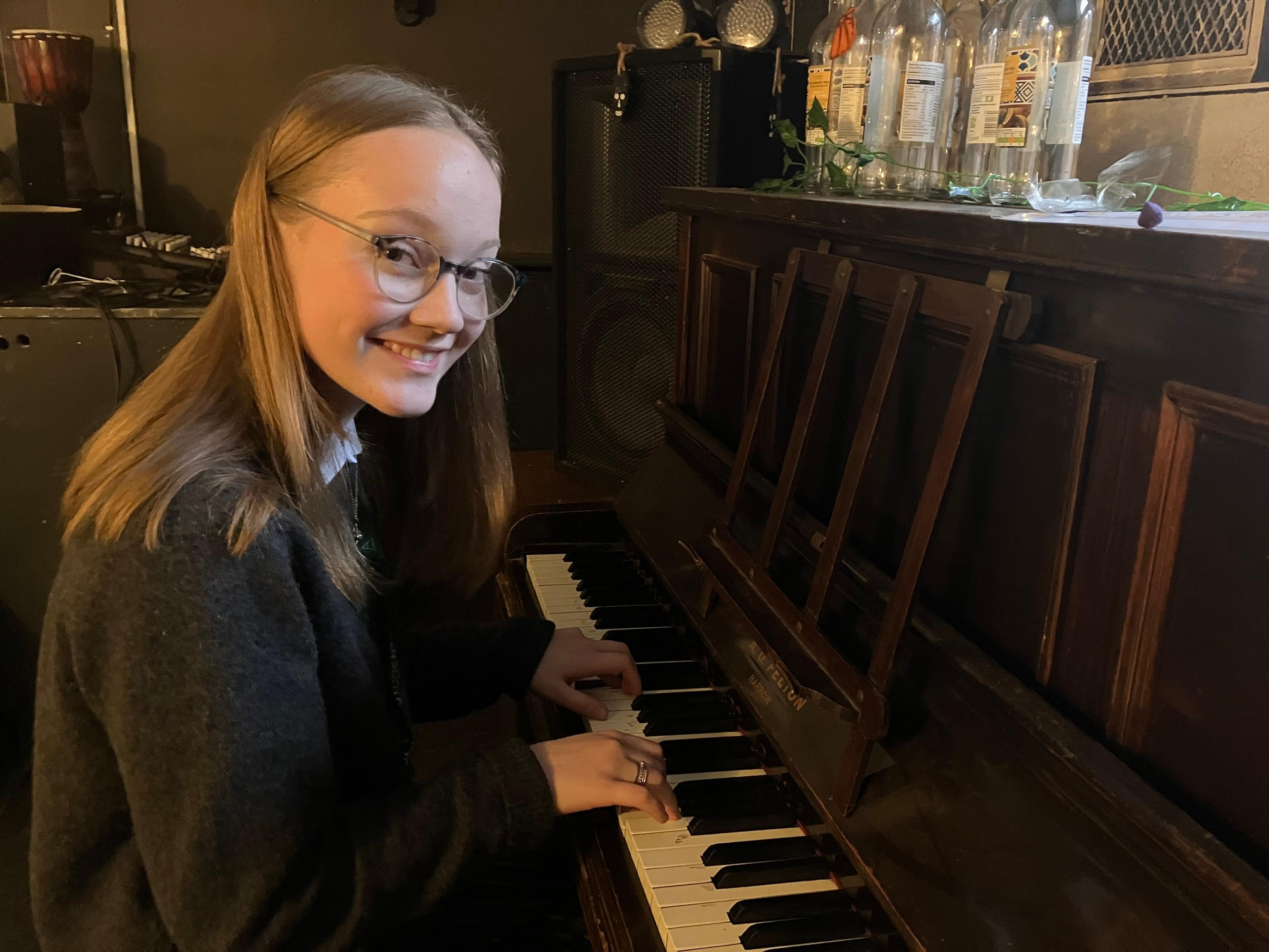 An image of a teenage girl playing a piano. She is smiling and is wearing glasses and has shoulder length brown hair.