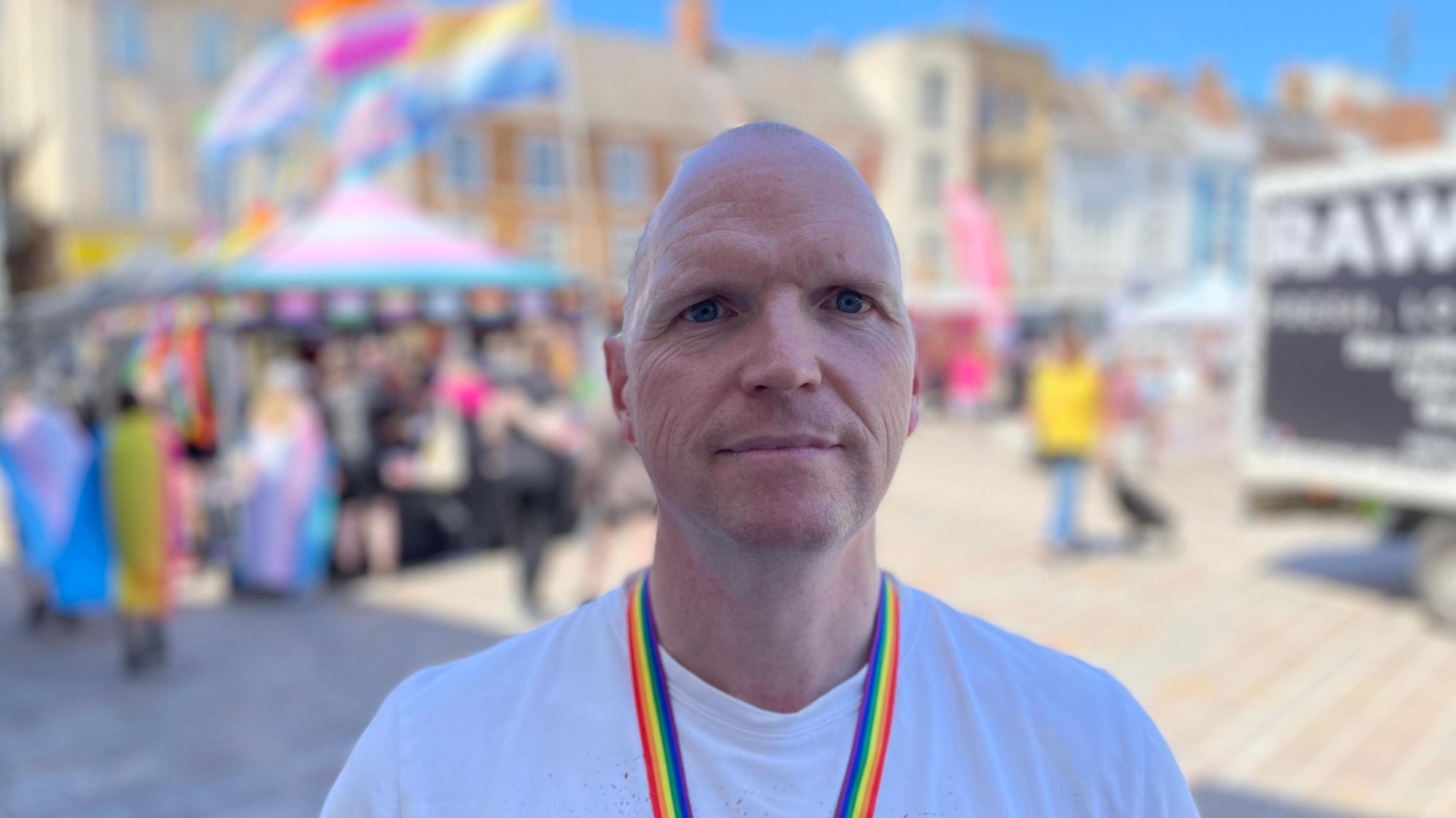 Nick is smiling at the camera while wearing a white top and a rainbow lanyard. He has blue eyes and no hair. Behind him people can be seen walking around with flags over their shoulders. 