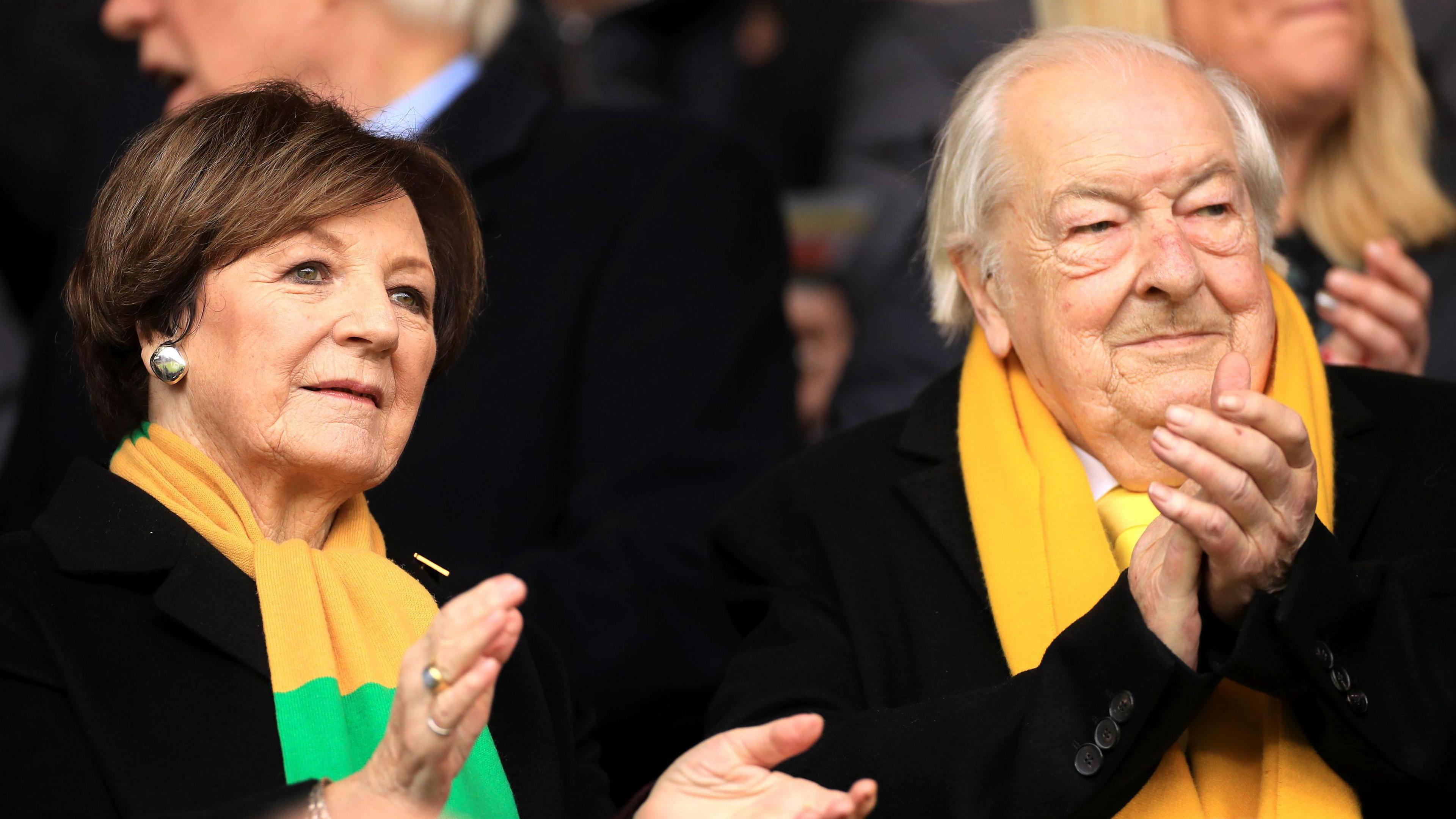 Delia Smith and Michael Wynn Jones cheer on Norwich City at Carrow Road in February 2023. They are both wearing black coats and yellow scarfs and are clapping.