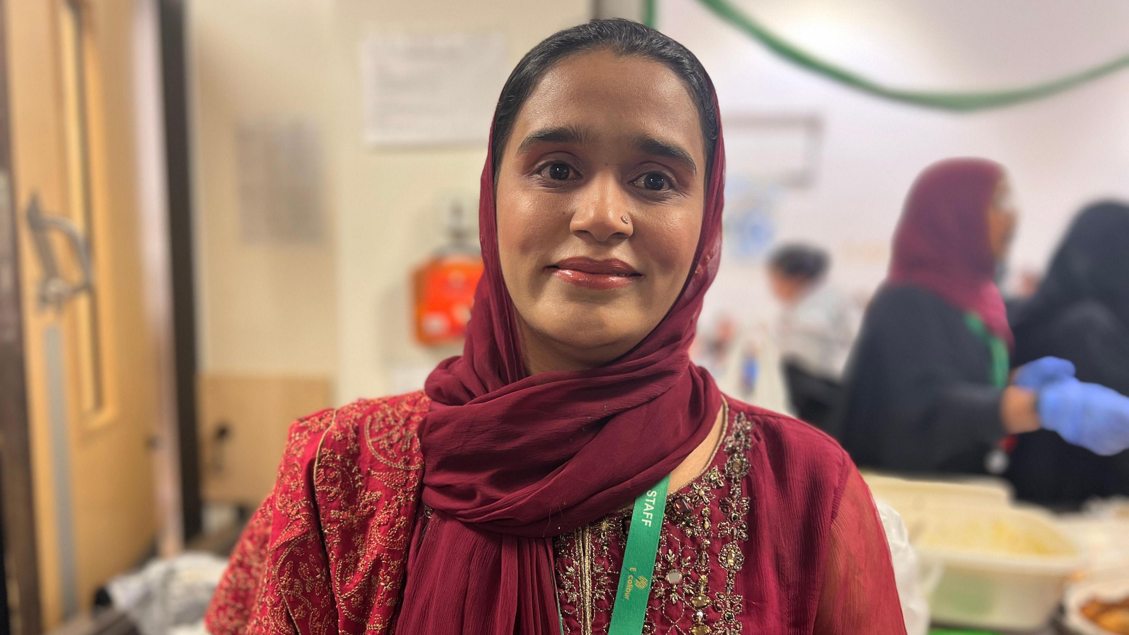 Ammara Ghazai, a woman wearing a deep red headscarf, smiling at the camera. She is wearing a green lanyard that says "staff". 