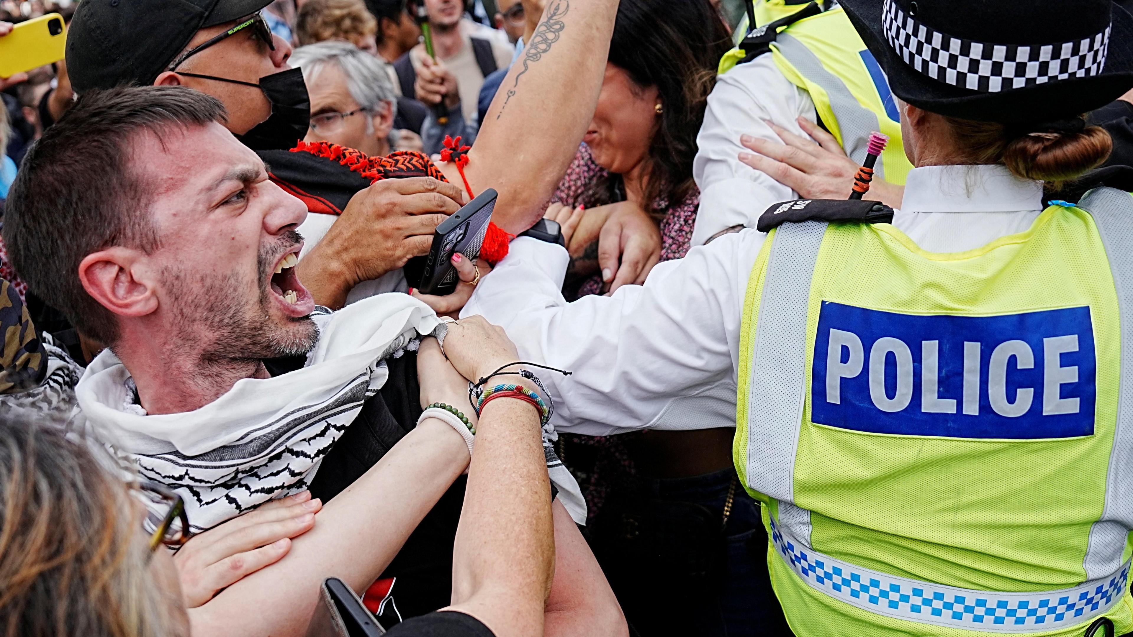 A man wearing a white and black patterned scarf stands amid a crowd looking at police officers who are confronting protesters. Several arms appear to be pulling him away
