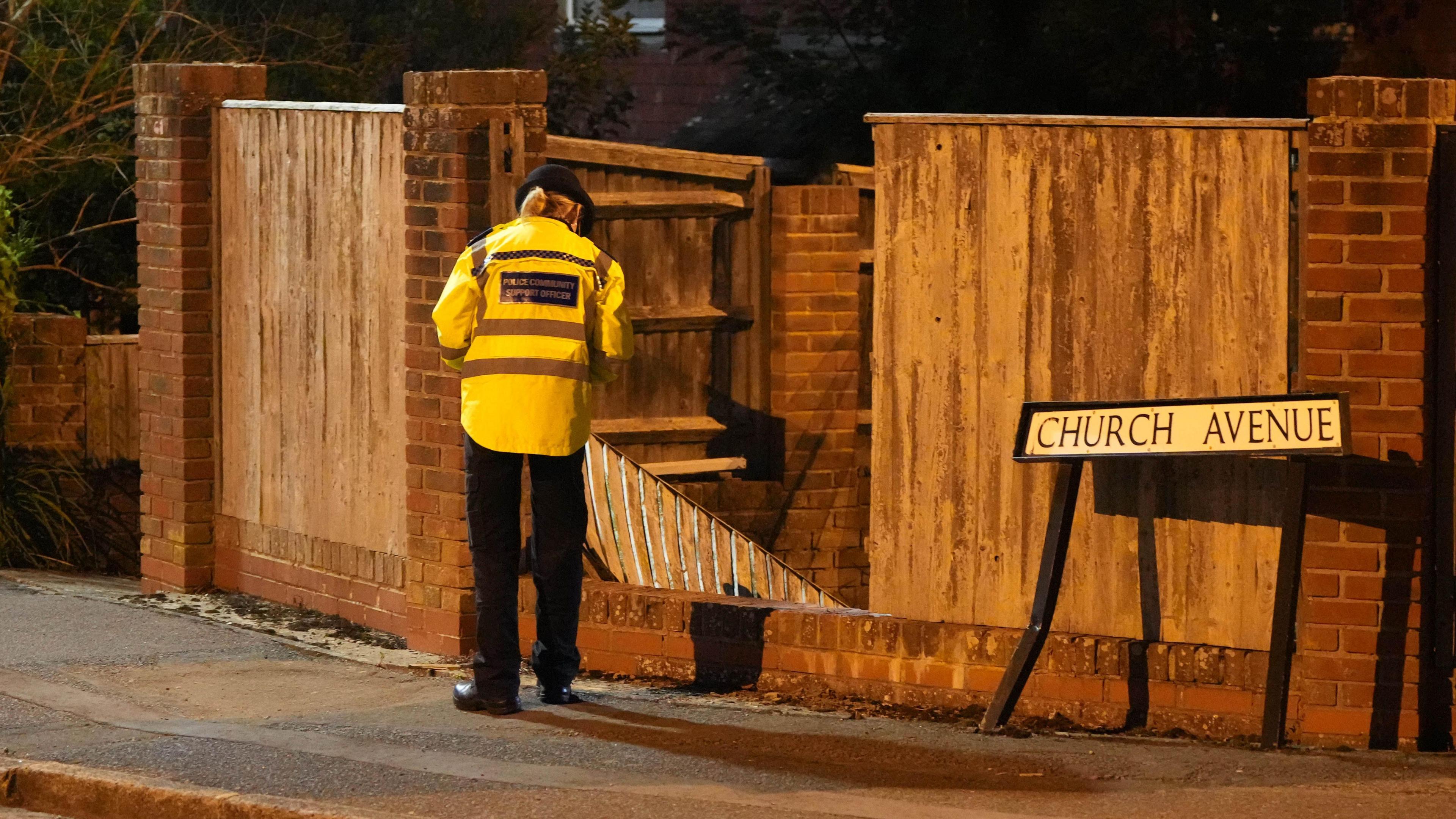 A police officer looking at a destroyed section of fencing. A road sign reading 'Church Avenue' is next to the fence and has also been damaged.
