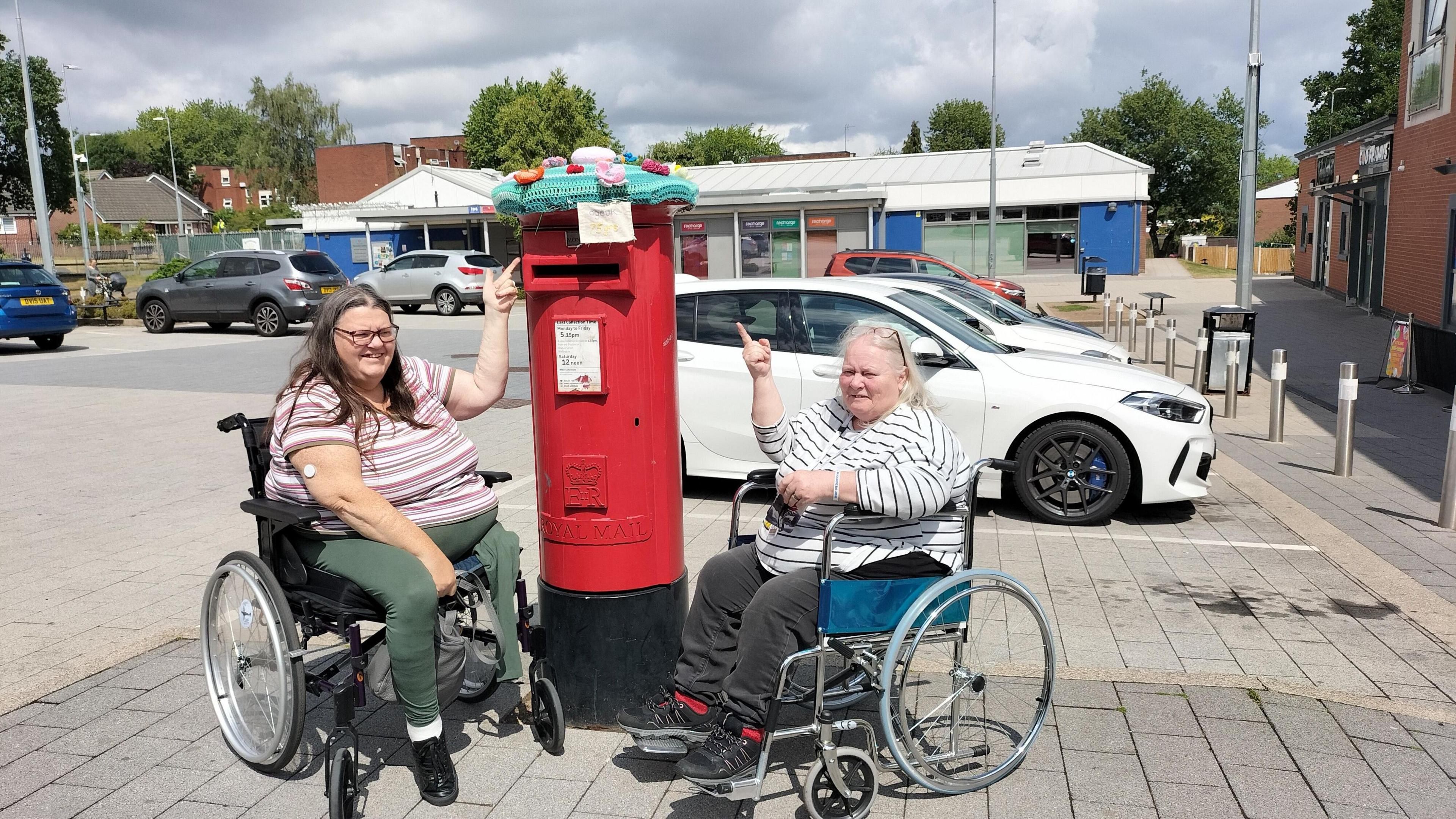 Two women in wheelchairs are next to a red Royal Mail post box. It has a topper on it that is knitted. It is teal blue with pink flowers on it. The women are pointing up at it.