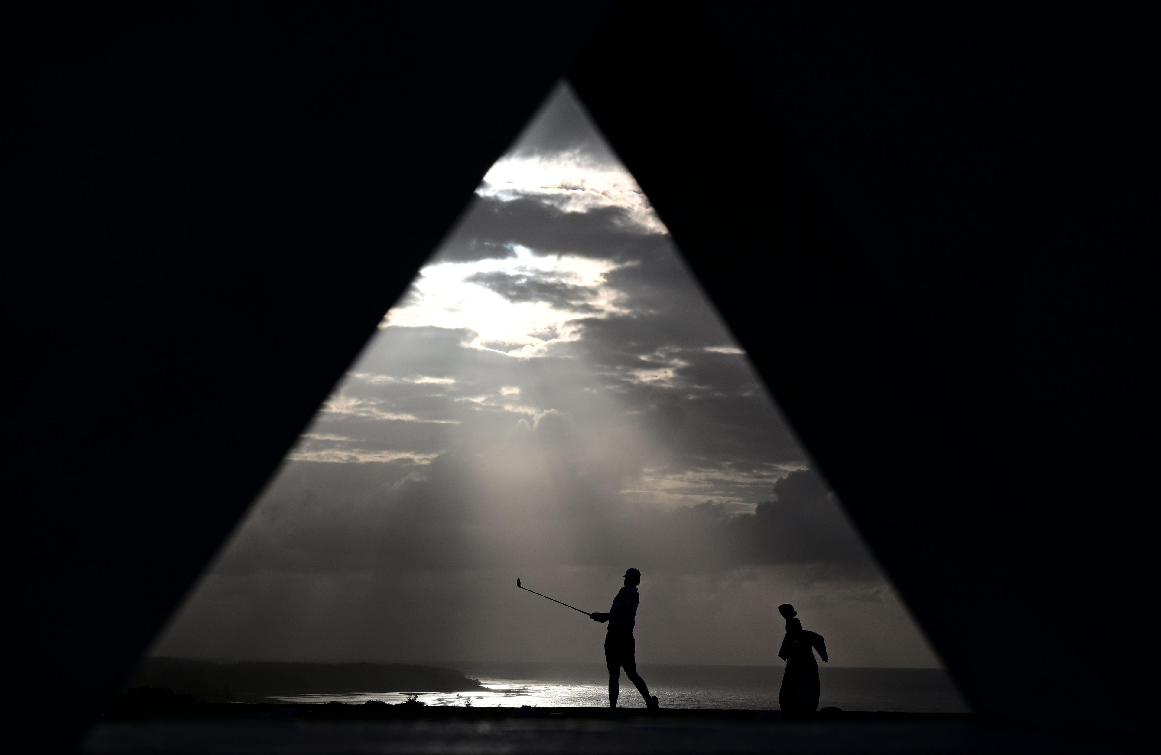 Silhouettes of two people are seen through a triangular opening, with one person holding a golf club as if preparing to swing. The background features a dramatic cloudy sky with rays of sunlight breaking through, illuminating the ocean horizon below. The scene is framed by dark geometric shapes, creating a striking contrast between the bright sky and the shadowed foreground.