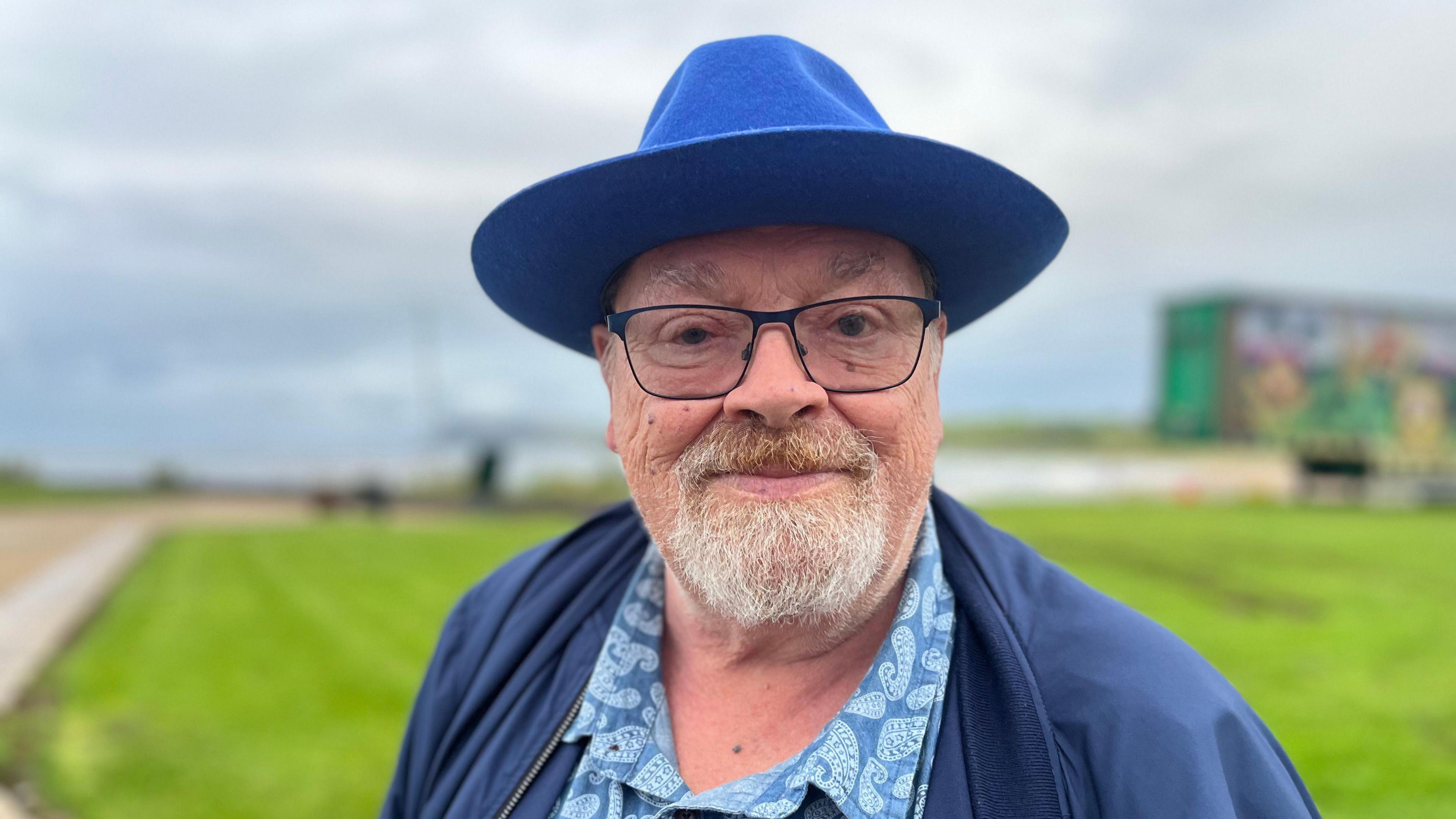 Older man wearing blue jacket and blue hat, with glasses and grey goatee, standing by the coast