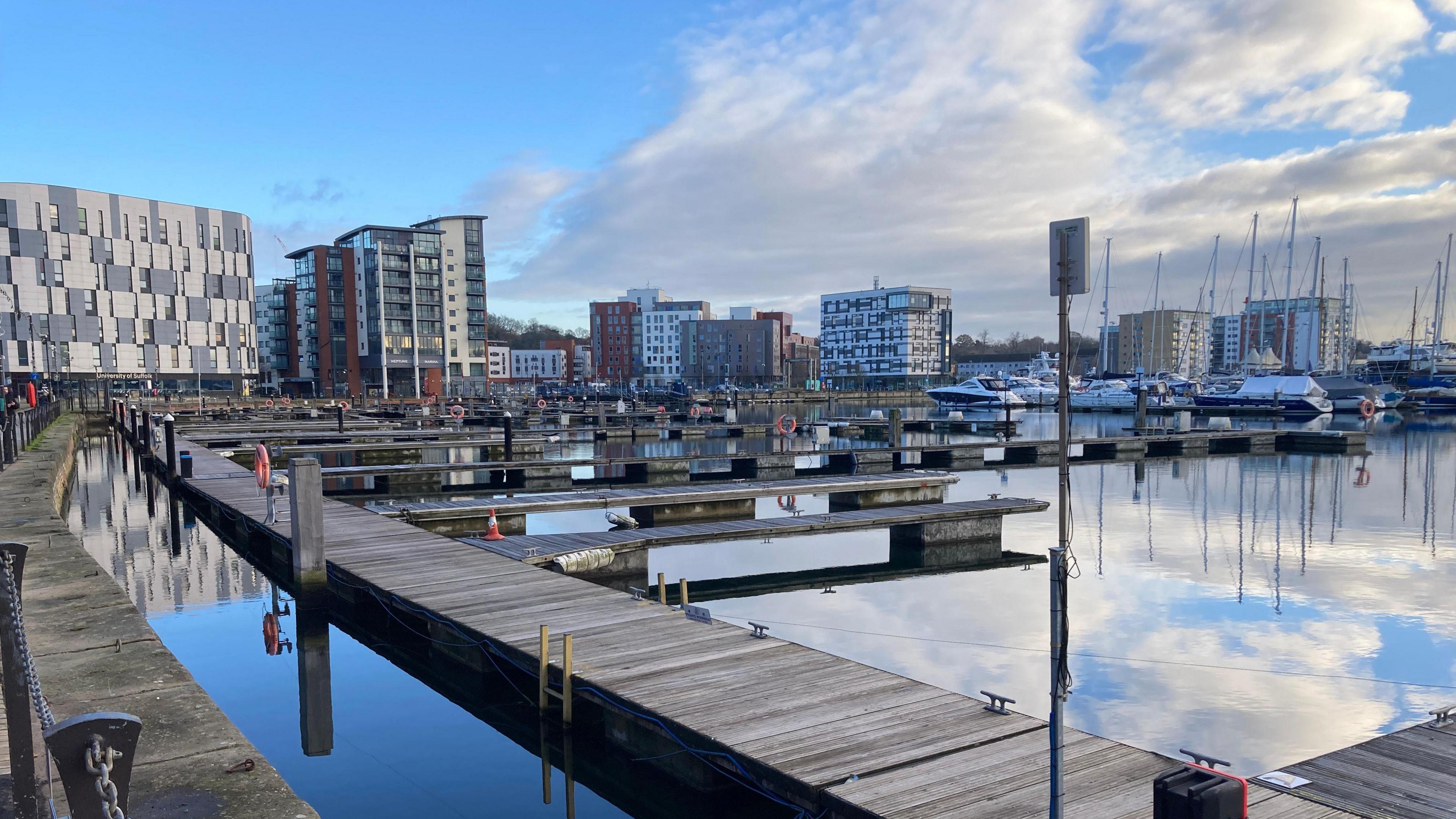 A view of Ipswich Marina. Boats can be seen resting on the water with empty moorings in the foreground. Large buildings can be seen in the distance.