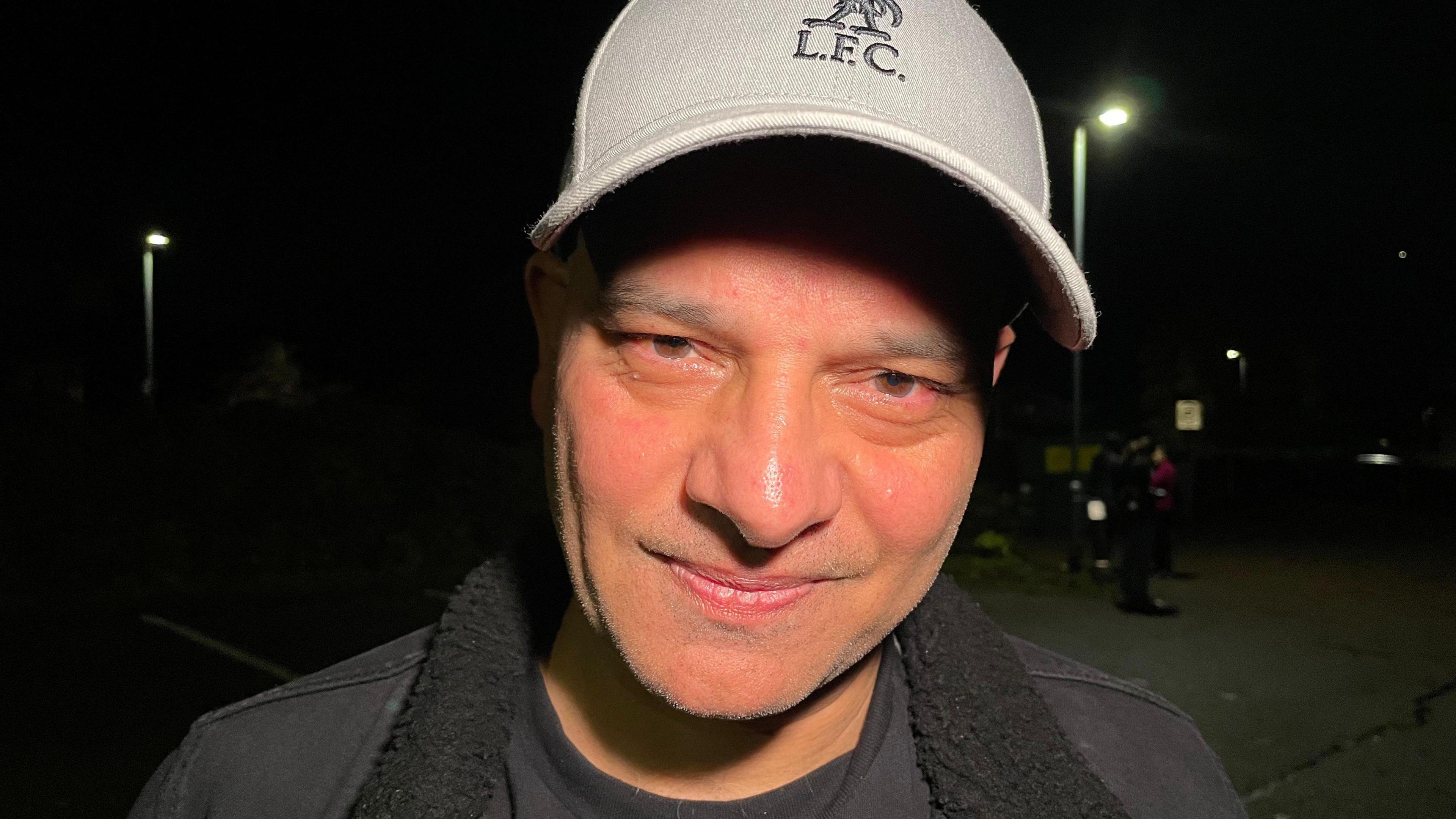 Shin Cheema is pictured at night. He is looking at the camera and wearing a Liverpool FC baseball cap. He is wearing a black t-shirt and black jacket with a black fleece collar. Streetlights can be seen behind him.