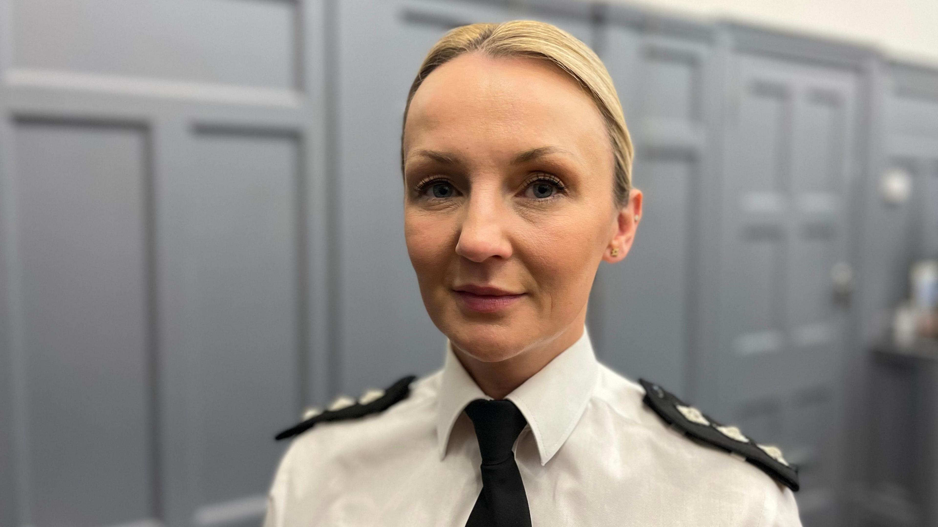 Chief Inspector Jennifer Busby is dressed in a police uniform wearing a white collared shirt with a black tie with shoulder pads.